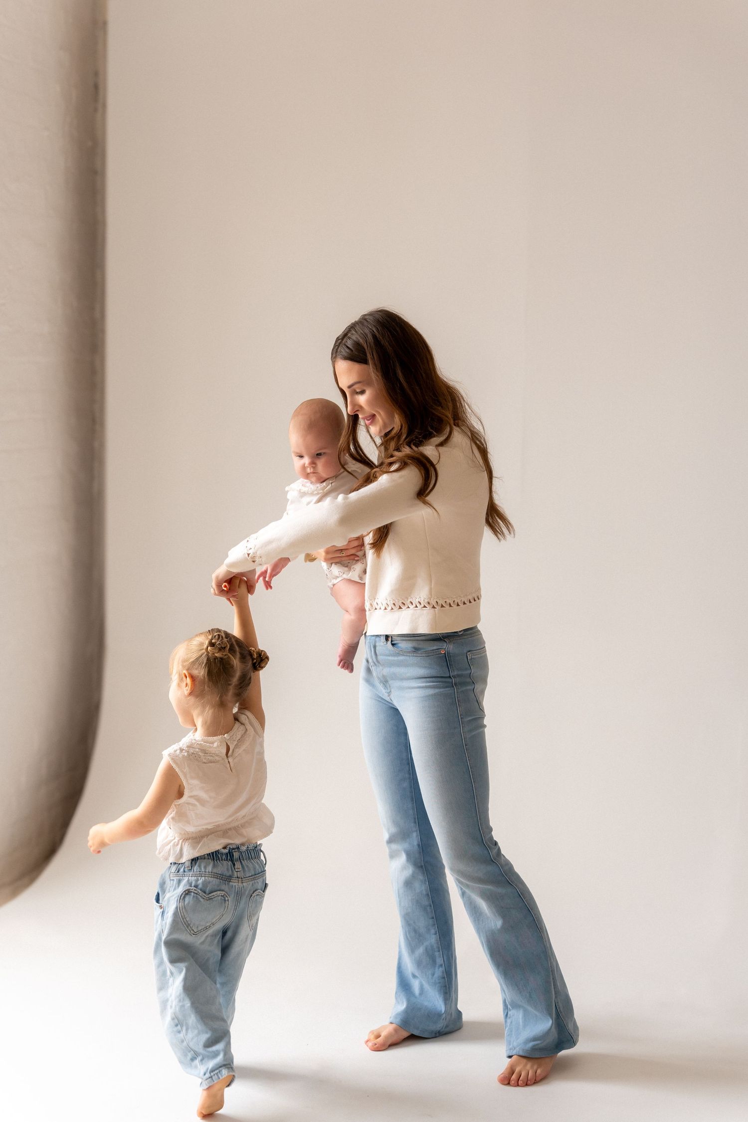 A mother in jeans and white sweater holds a baby while her toddler daughter reaches up to her hand in a studio.