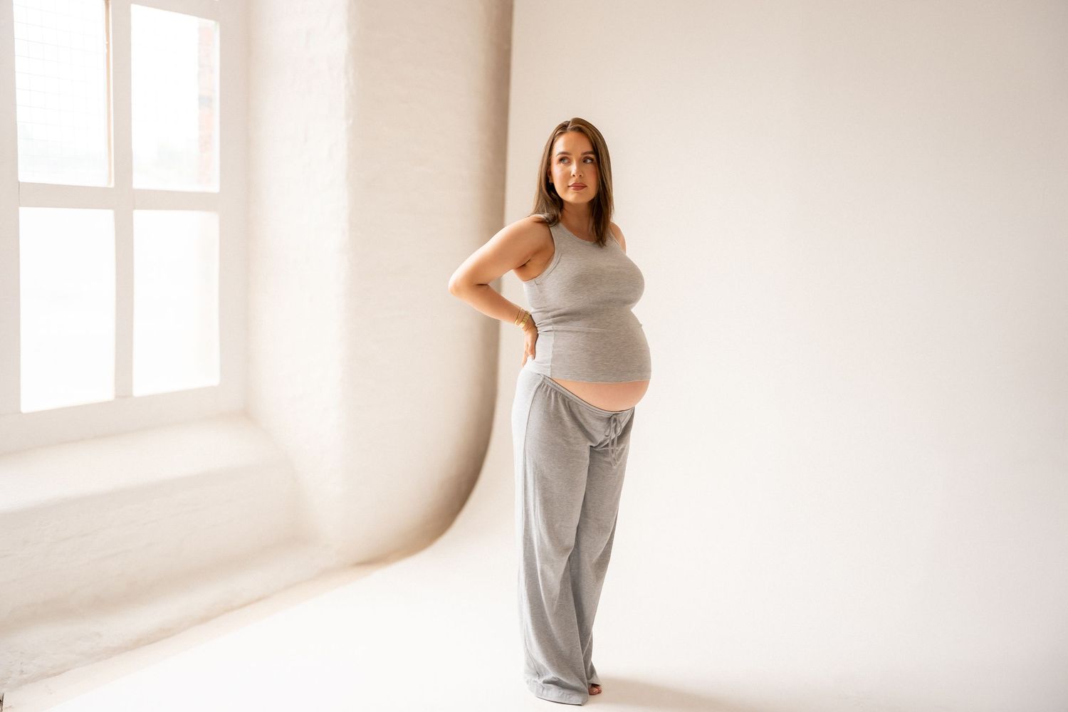 Pregnant woman in gray crop top and pants posing in bright white studio, showcasing baby bump.