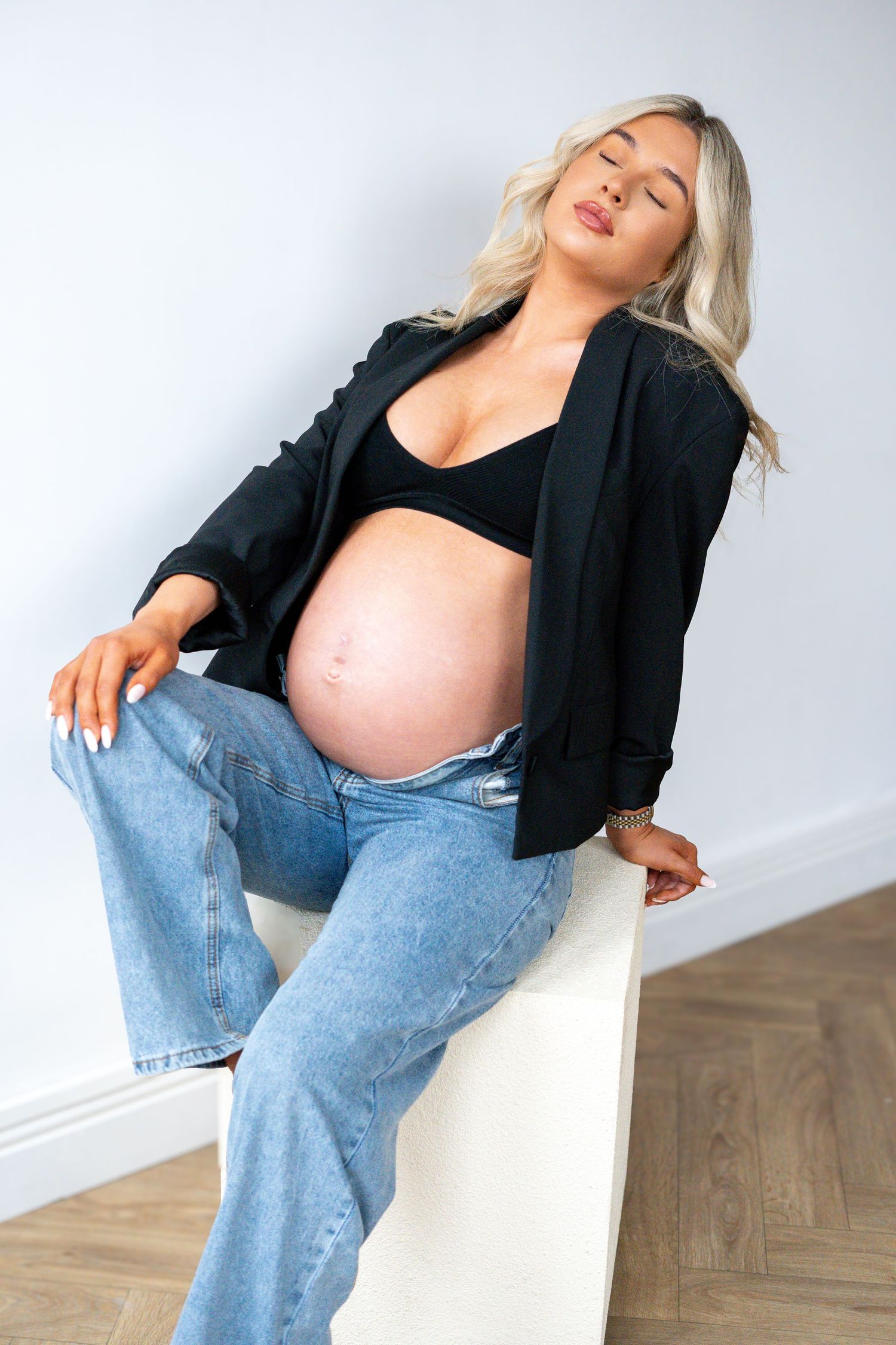 Pregnant woman in black blazer and blue jeans sitting on white cube with eyes closed, bare bump showing.