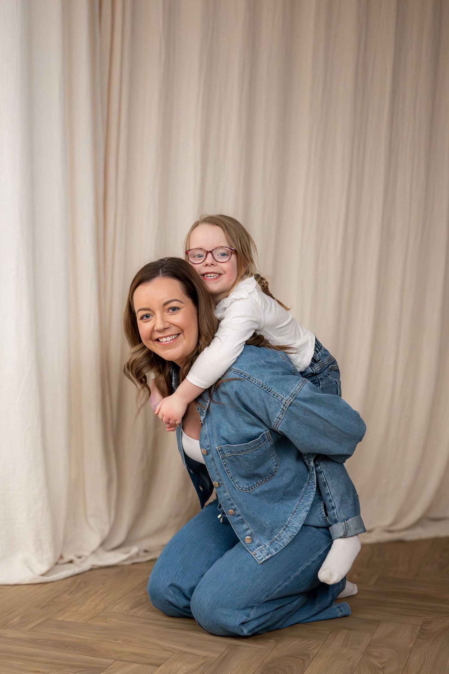 A smiling mother gives her young daughter with glasses a piggyback ride indoors.