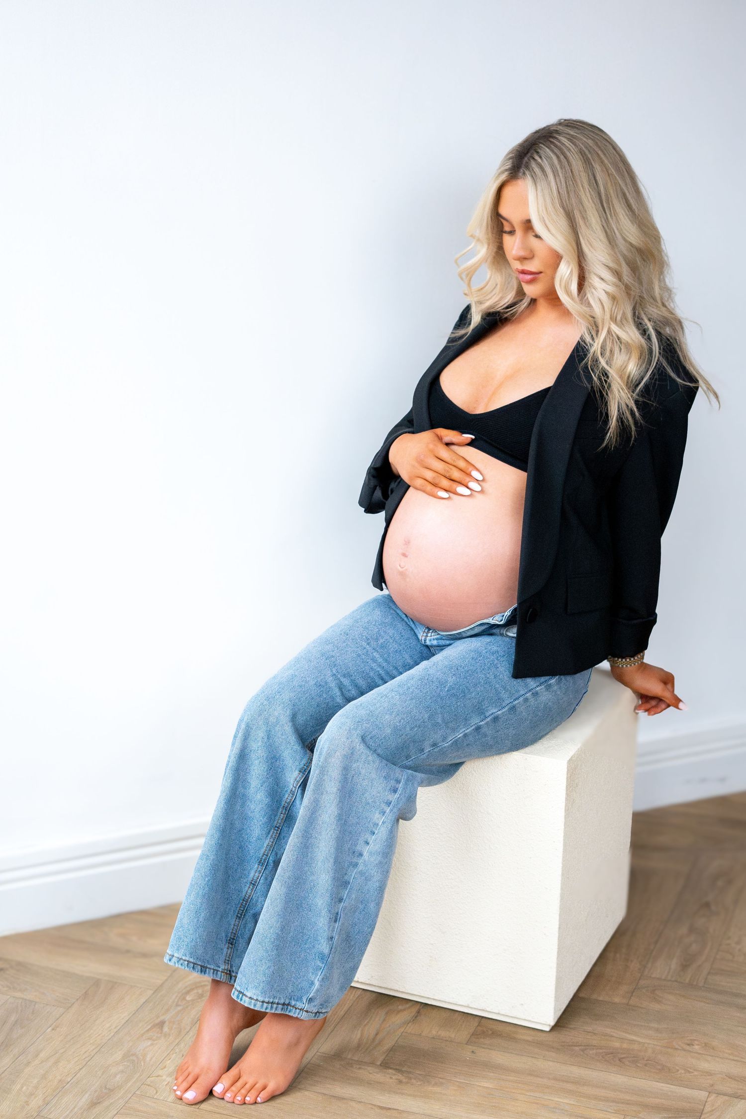 Pregnant woman in black blazer and jeans sitting on white cube, looking down at bare baby bump.