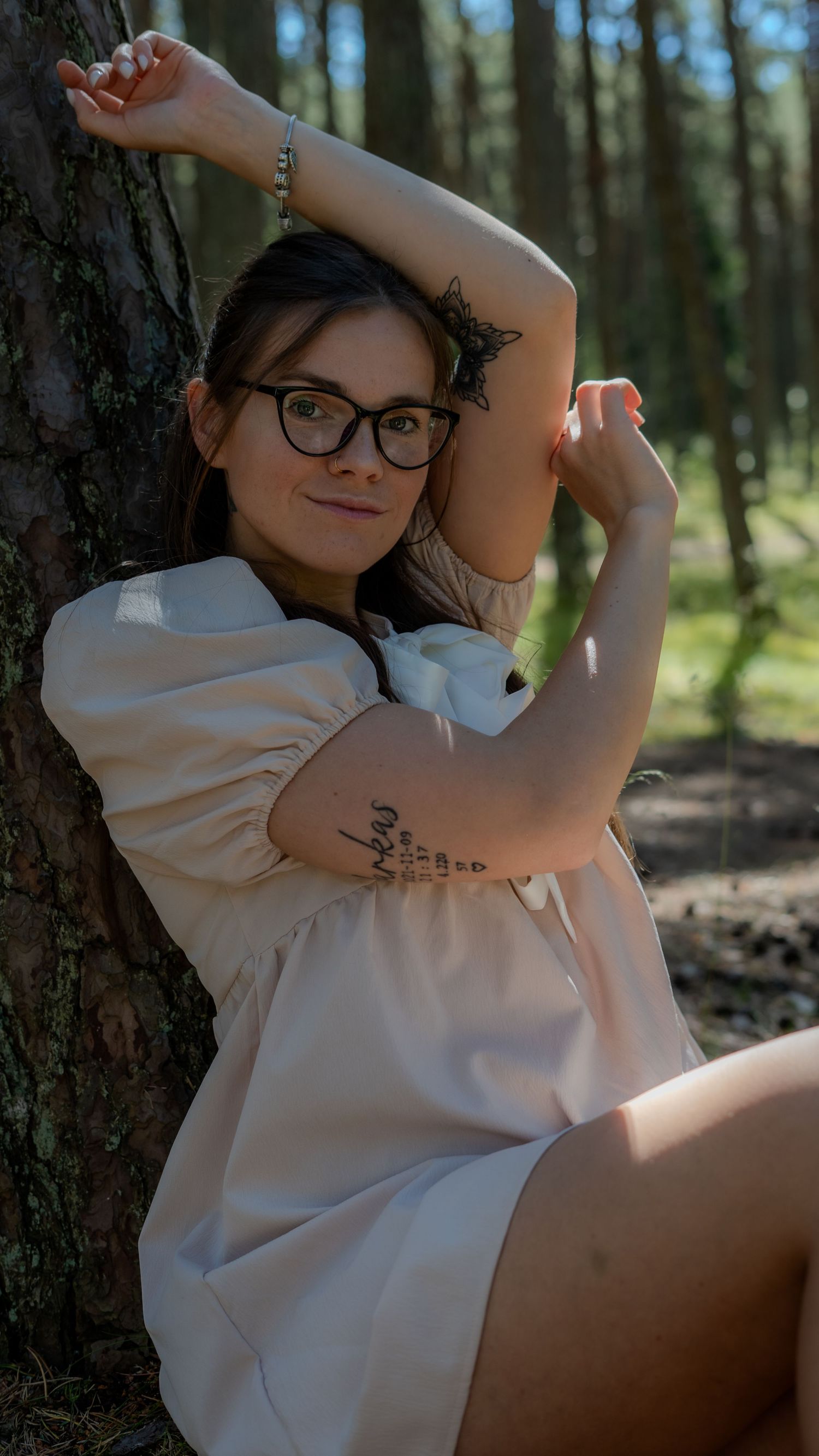 Young woman with glasses and tattoos in a beige dress poses against a pine tree in a sunlit forest.