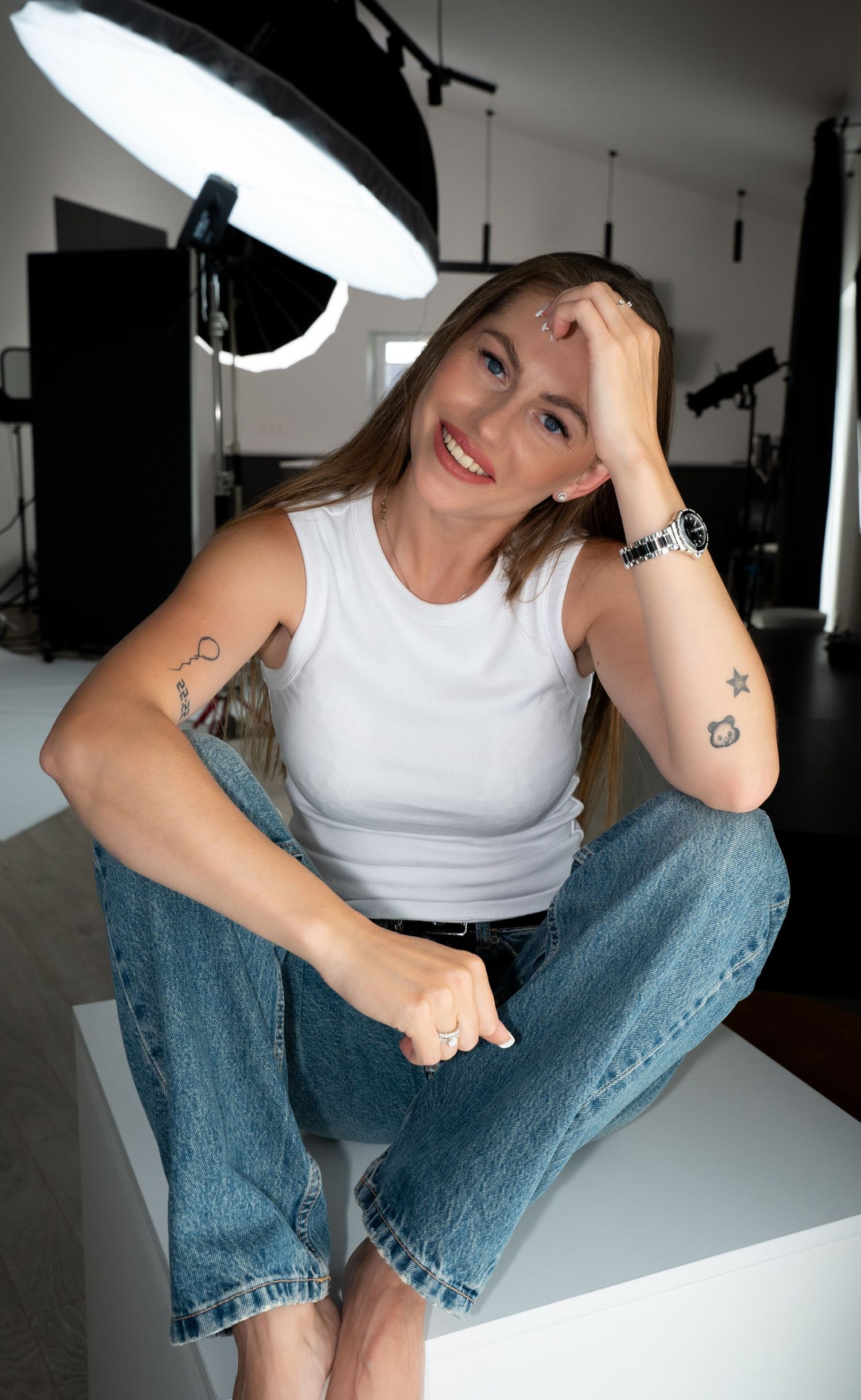 Smiling tattooed blonde woman in white tank top and jeans sitting cross-legged under studio ring light.