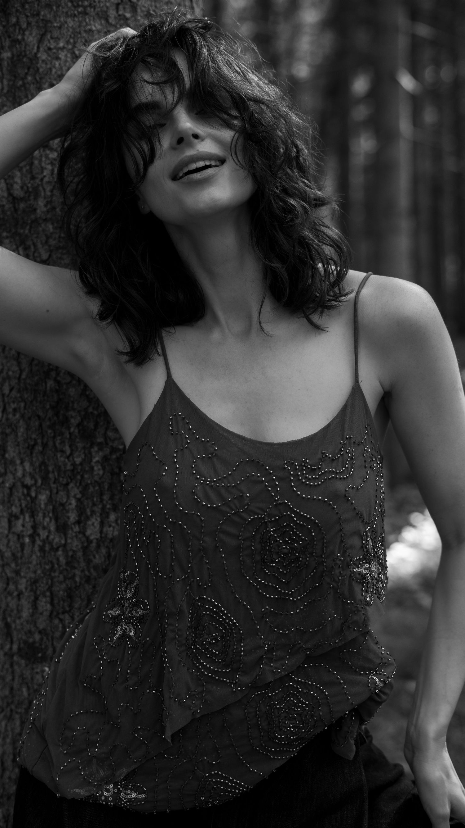 Black and white photo of woman in beaded camisole leaning against tree with hand in wavy hair.