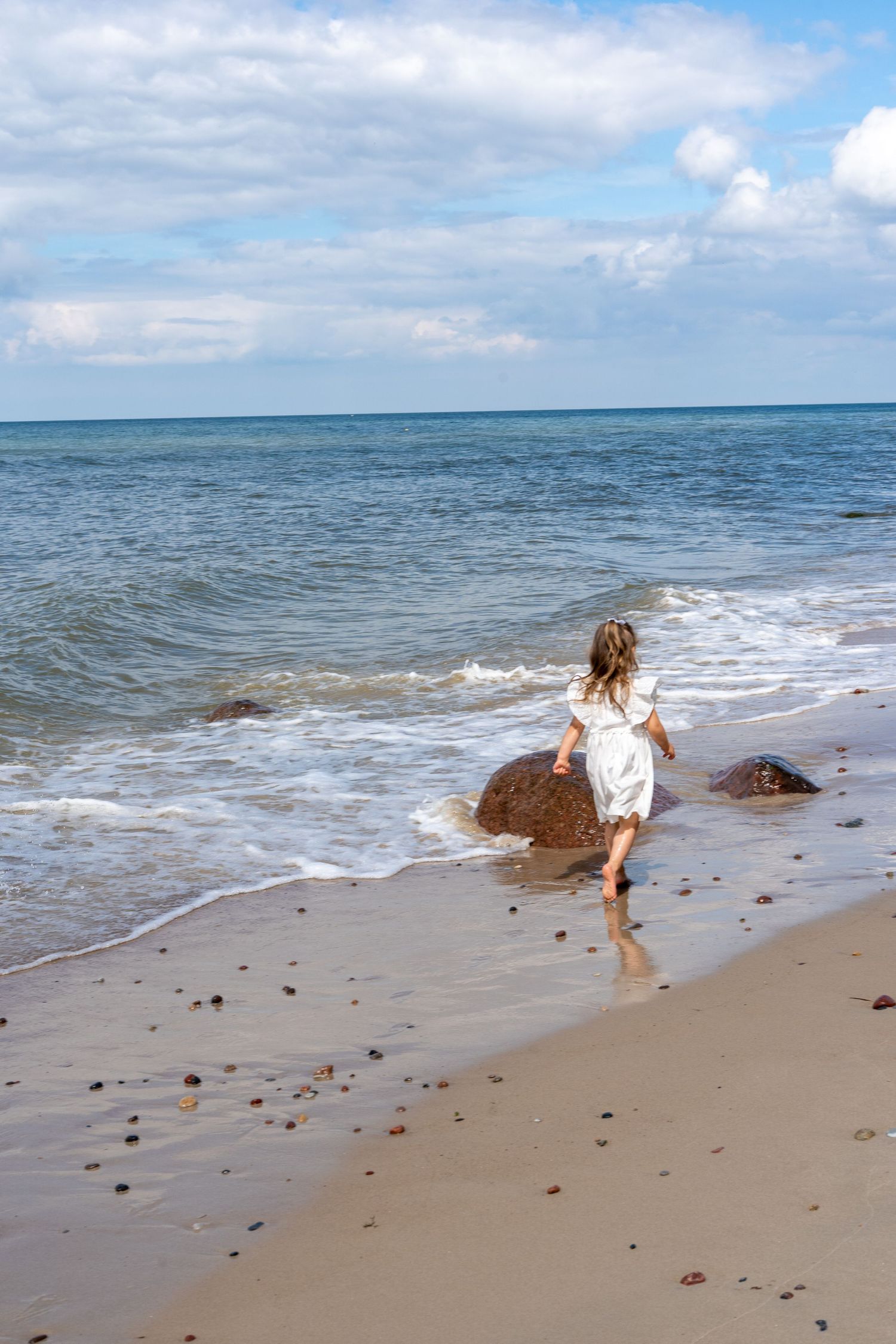 Young girl in a white dress runs barefoot along a sandy beach toward ocean waves.