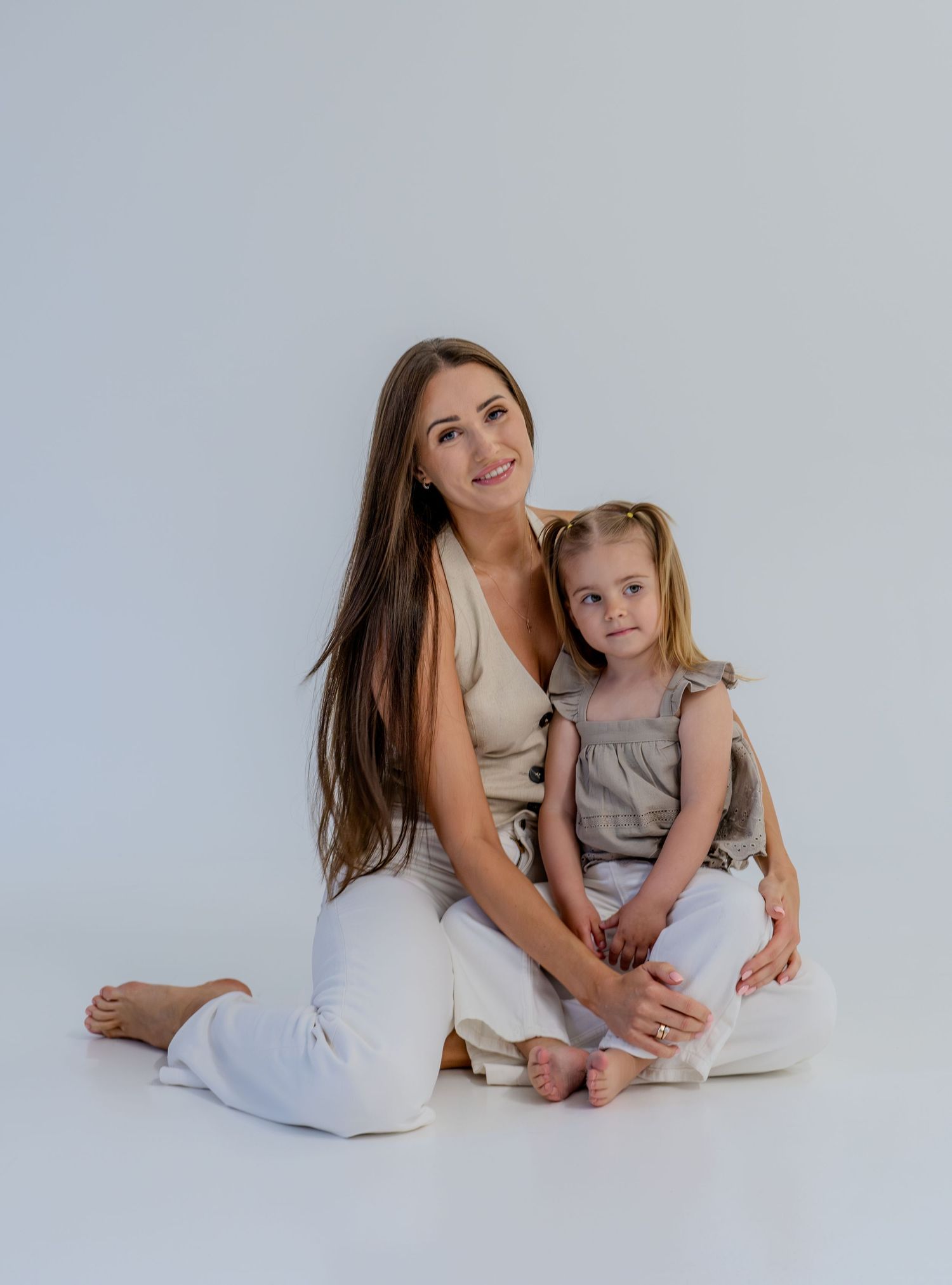 A brunette mother sits on the floor with her young blonde daughter, both wearing white pants in a studio.