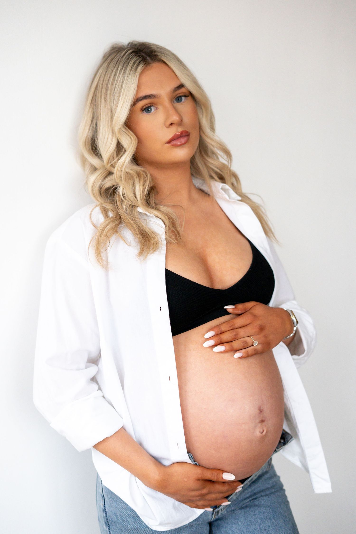 Pregnant blonde woman in open white shirt and black bralette gently holding baby bump.