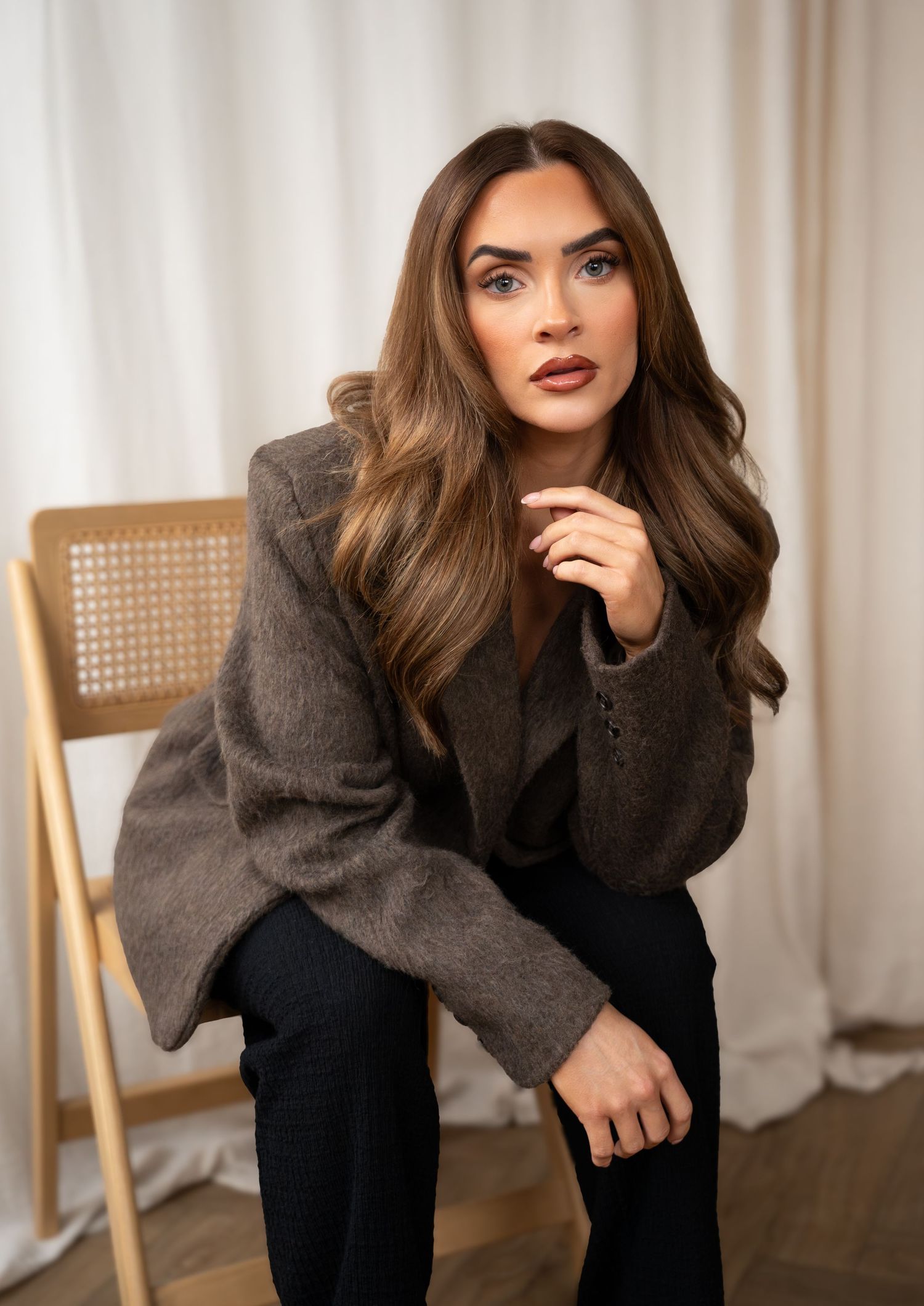 Brunette woman in gray blazer posing on wooden chair with curtain backdrop.