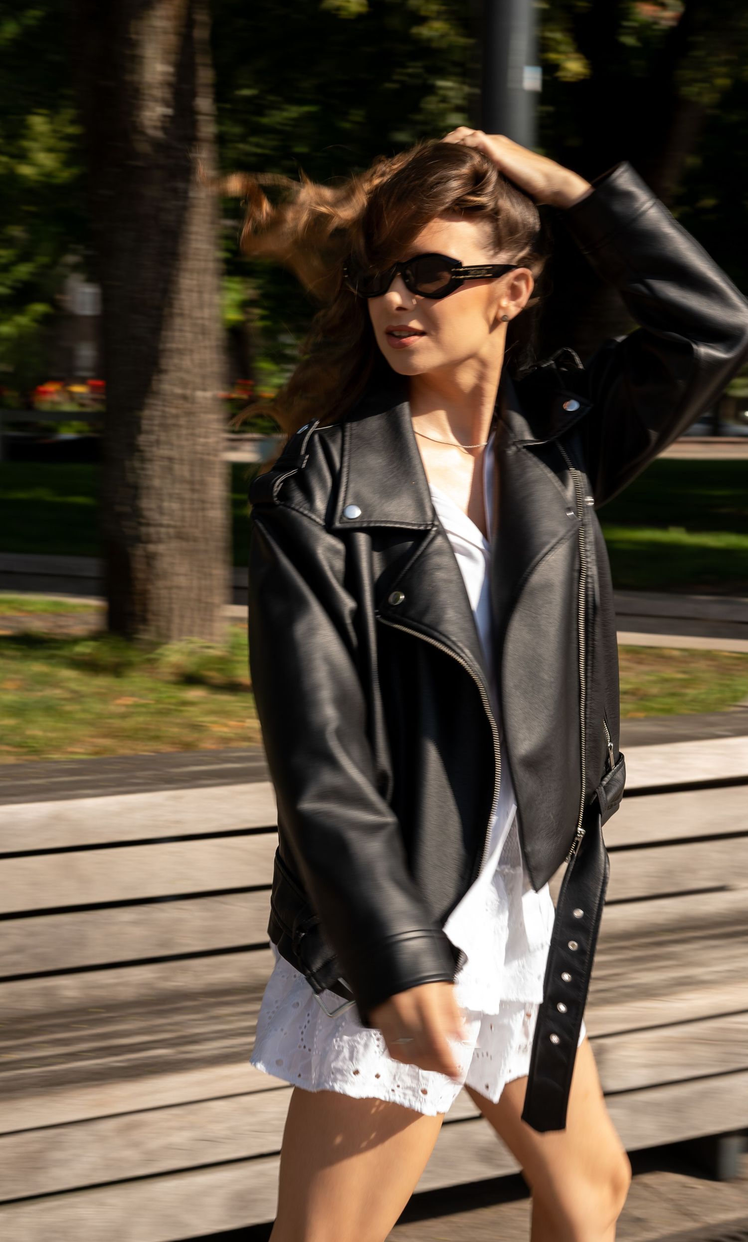 Woman in black leather biker jacket and white mini dress walking in sunny park with sunglasses on.