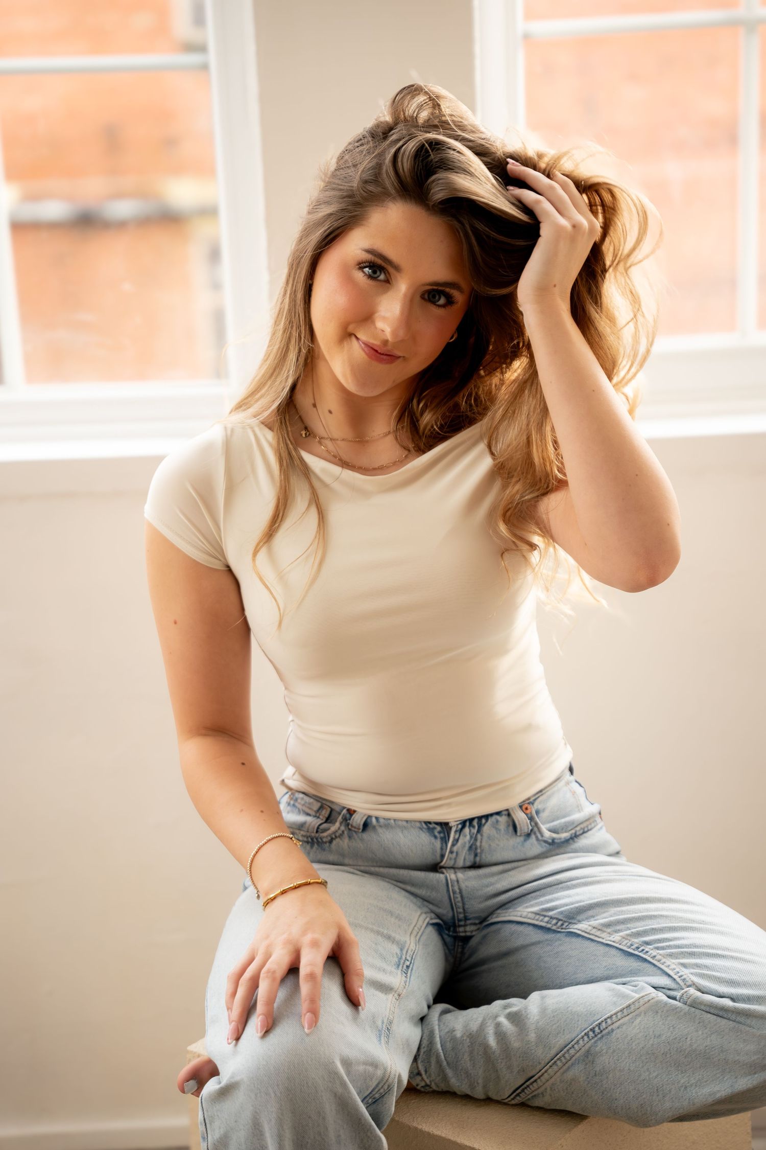 Smiling woman in white t-shirt and ripped jeans sits by a window running fingers through her hair.