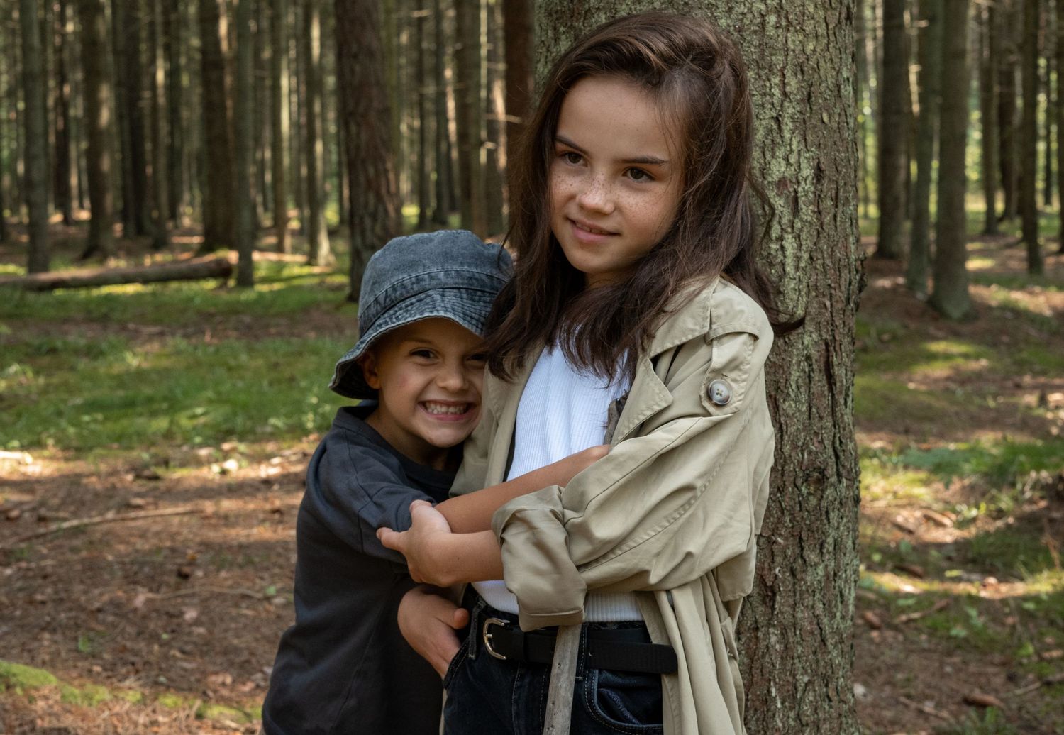Two children, a girl in a trench coat and a boy in a hat, embrace while leaning against a tree in the woods.