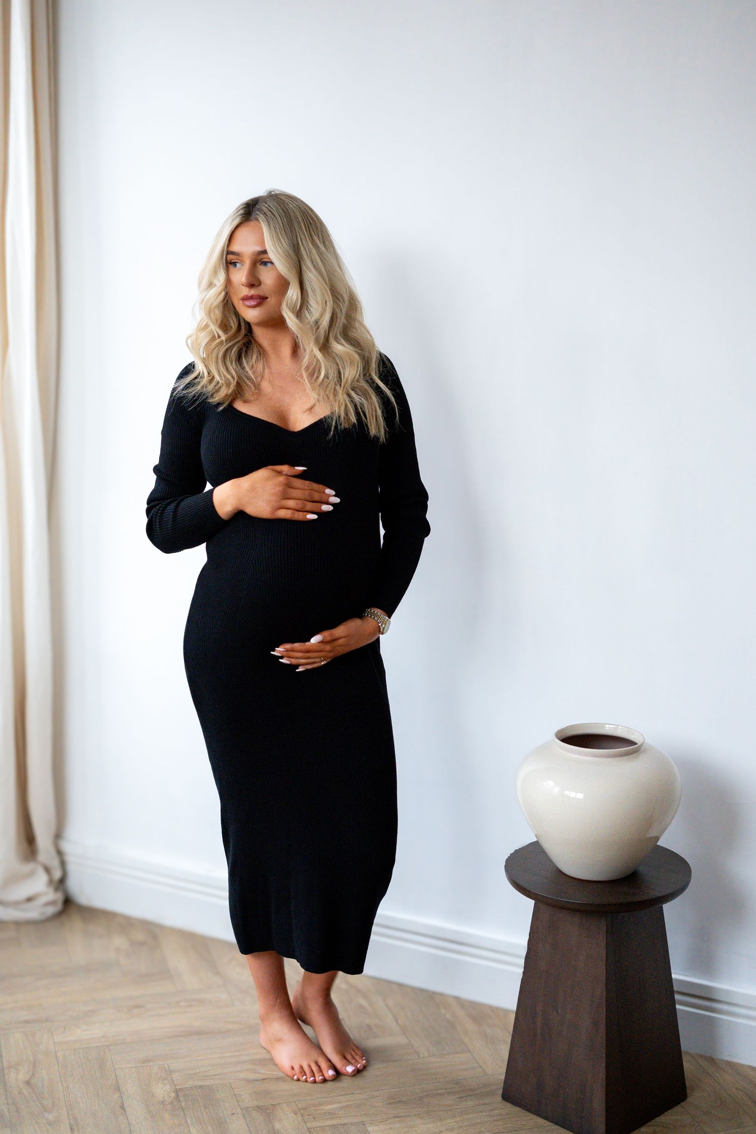 Pregnant woman in black ribbed midi dress holding baby bump, standing barefoot next to decorative vase on dark stool.
