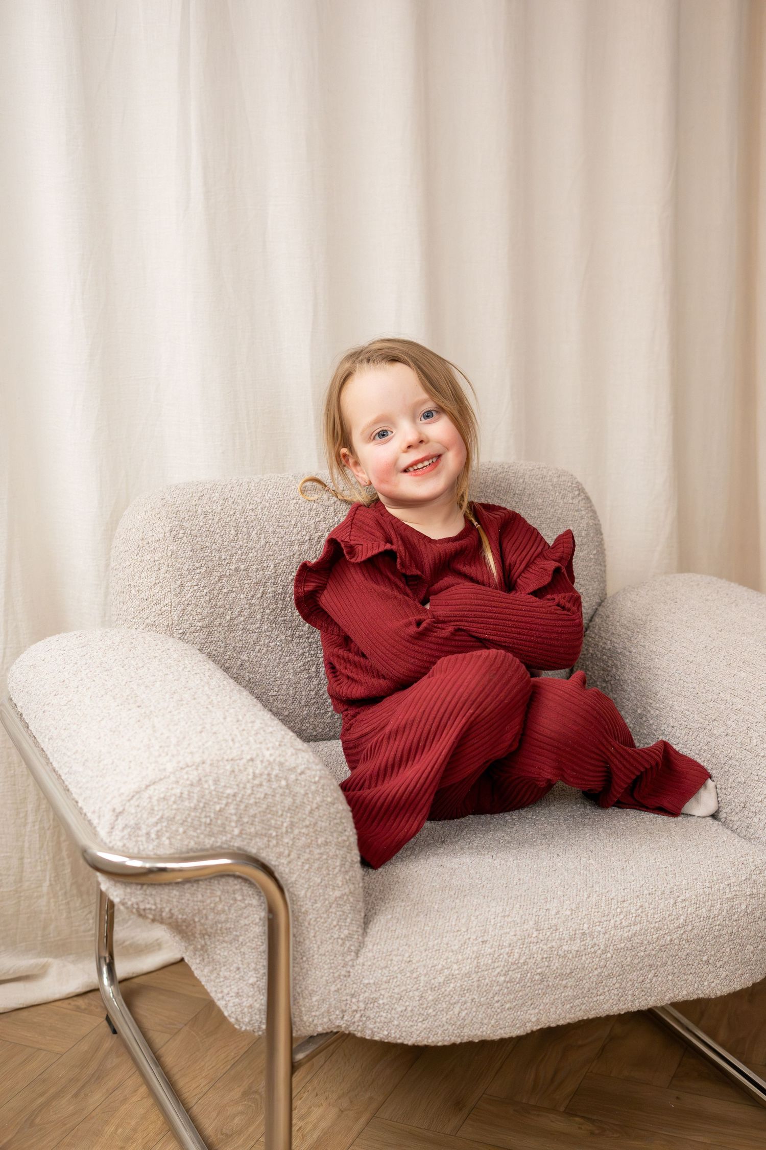 A cheerful young girl in a burgundy outfit sits smiling in a cozy white boucle armchair.