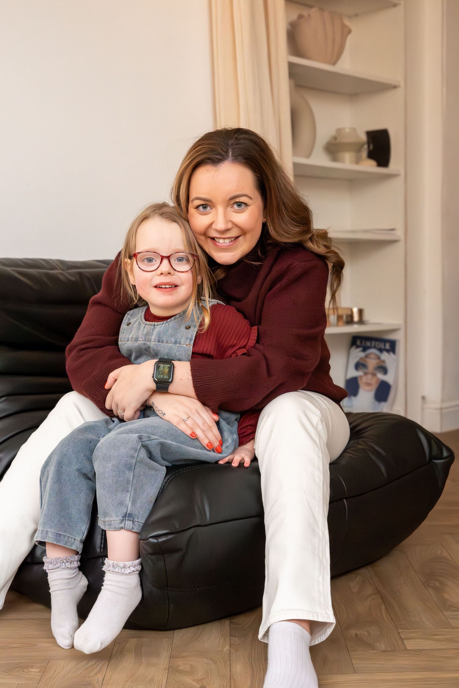 Smiling brunette mother hugging young daughter in glasses and denim overalls on black bean bag chair.