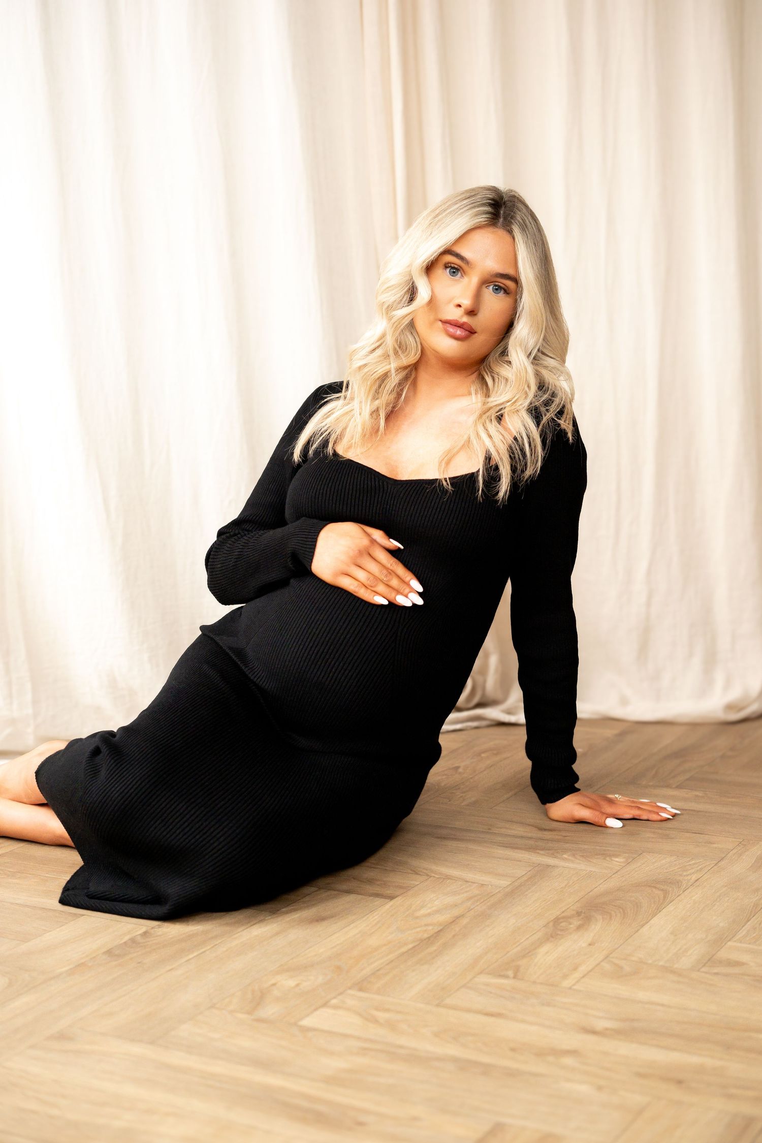 Pregnant woman in black dress sitting gracefully on wooden floor cradling baby bump in bright studio.