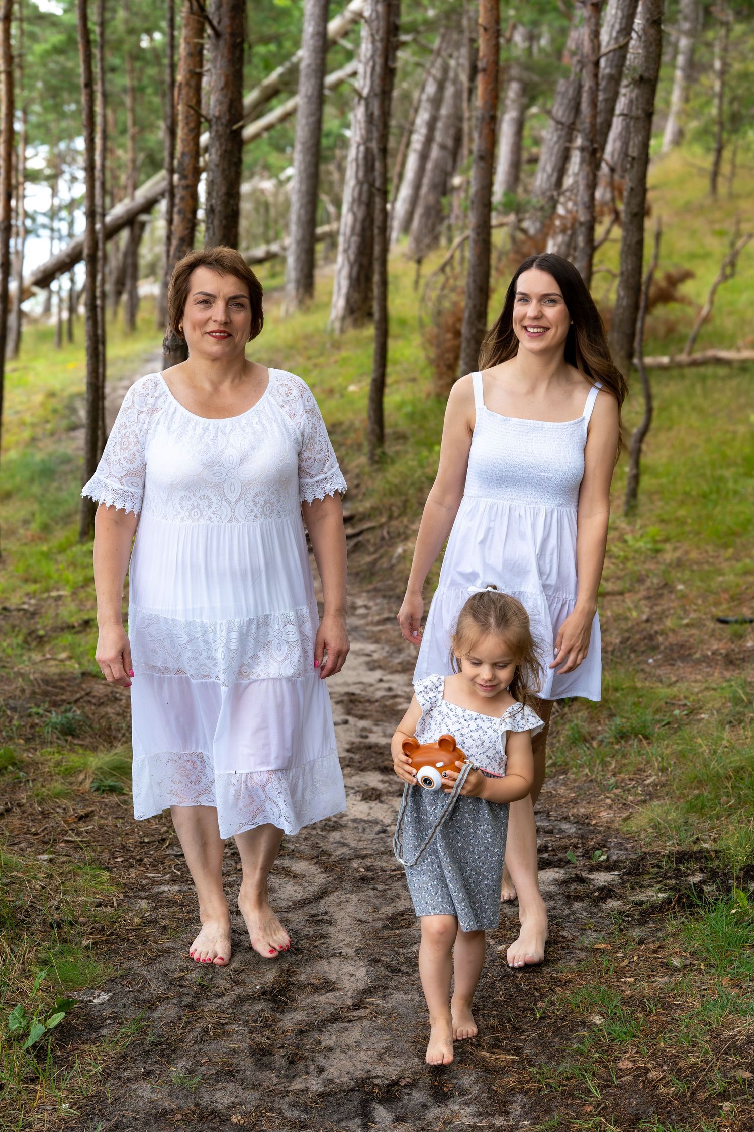 Three generations of women in white dresses walk barefoot on a forest path among pine trees.