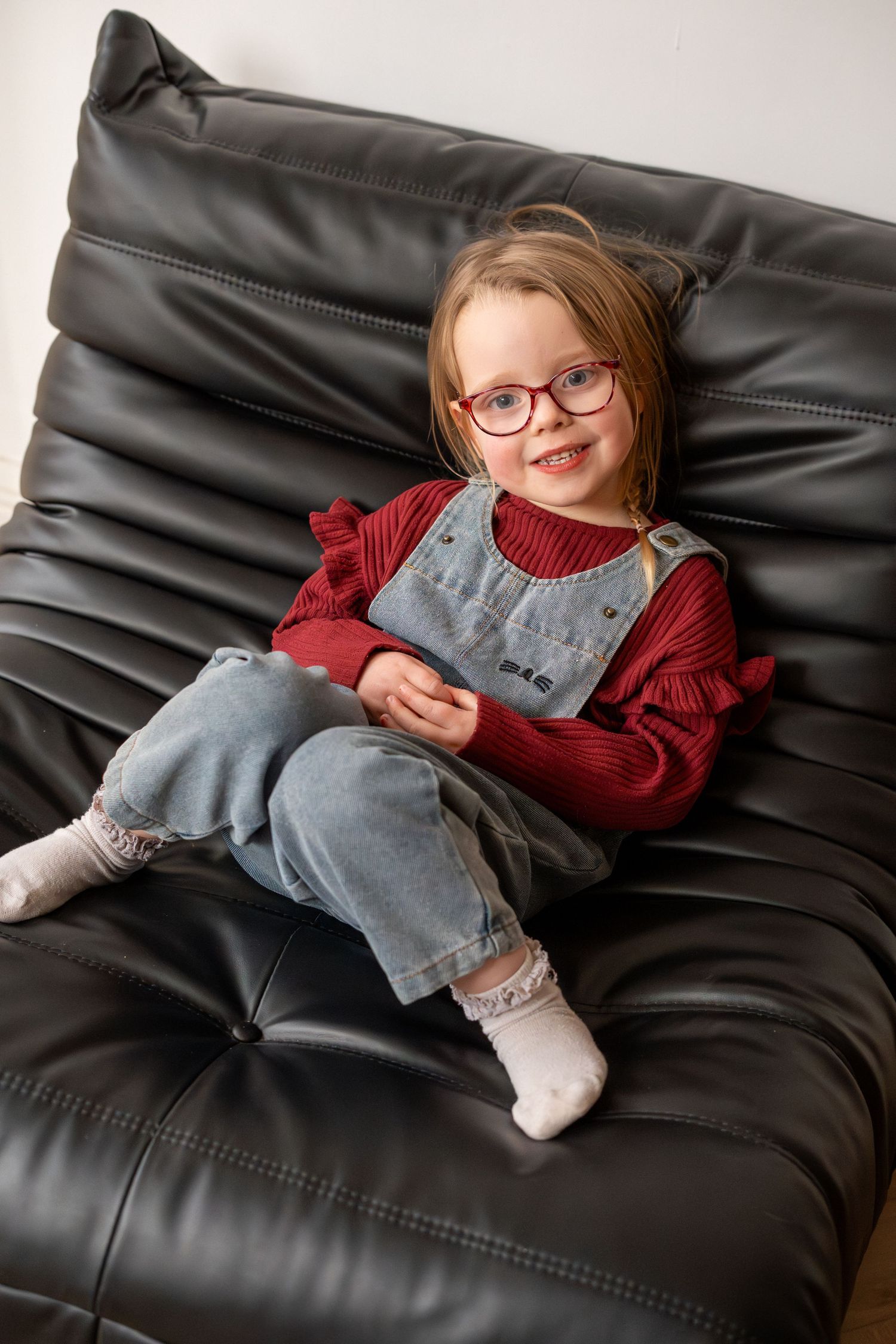 A cute young girl wearing glasses and denim overalls smiles while sitting on a black leather chair.