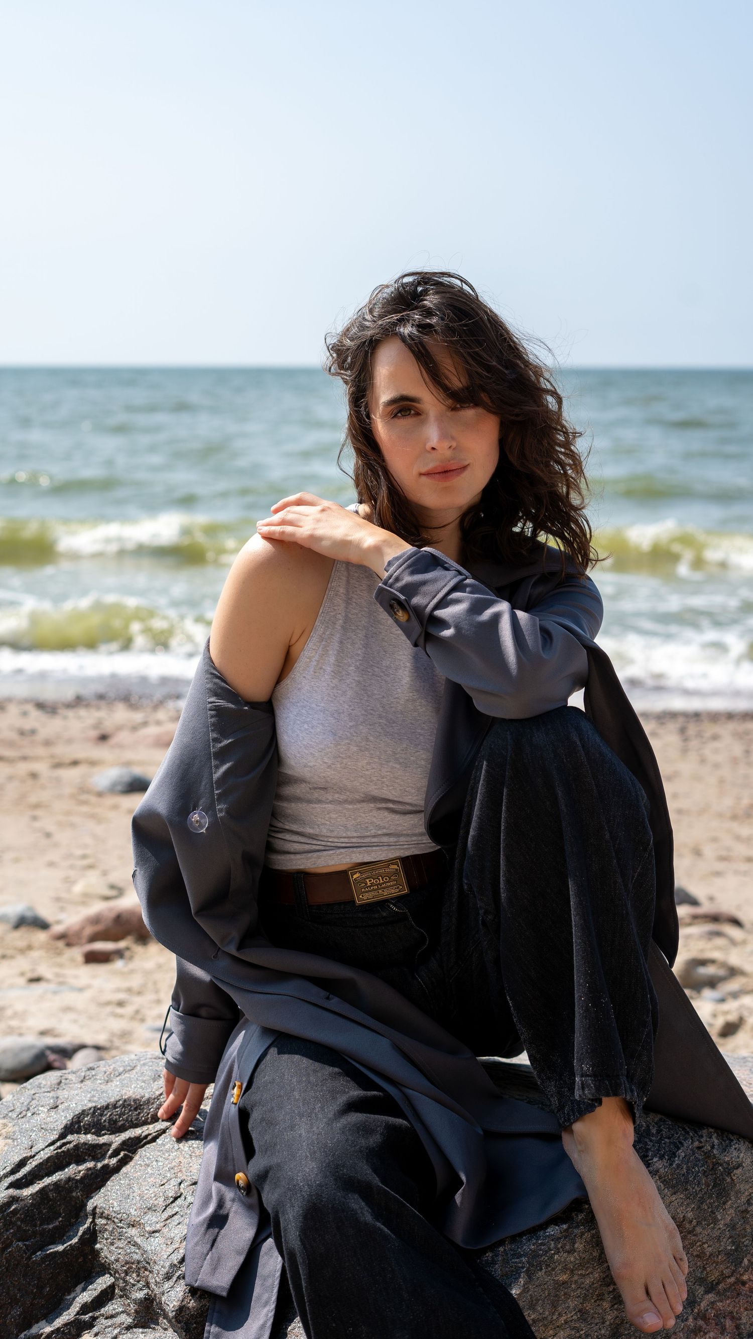 Woman in gray tank top and dark jacket sitting on beach rocks with ocean waves in background.