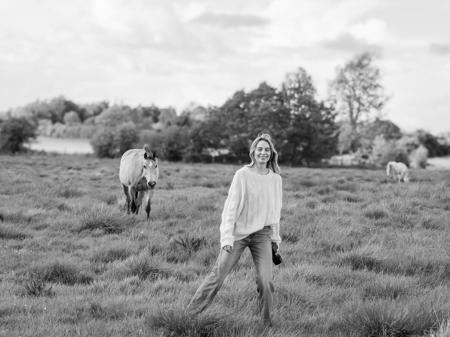Black and white photo of blonde woman standing in field with horses grazing behind her.