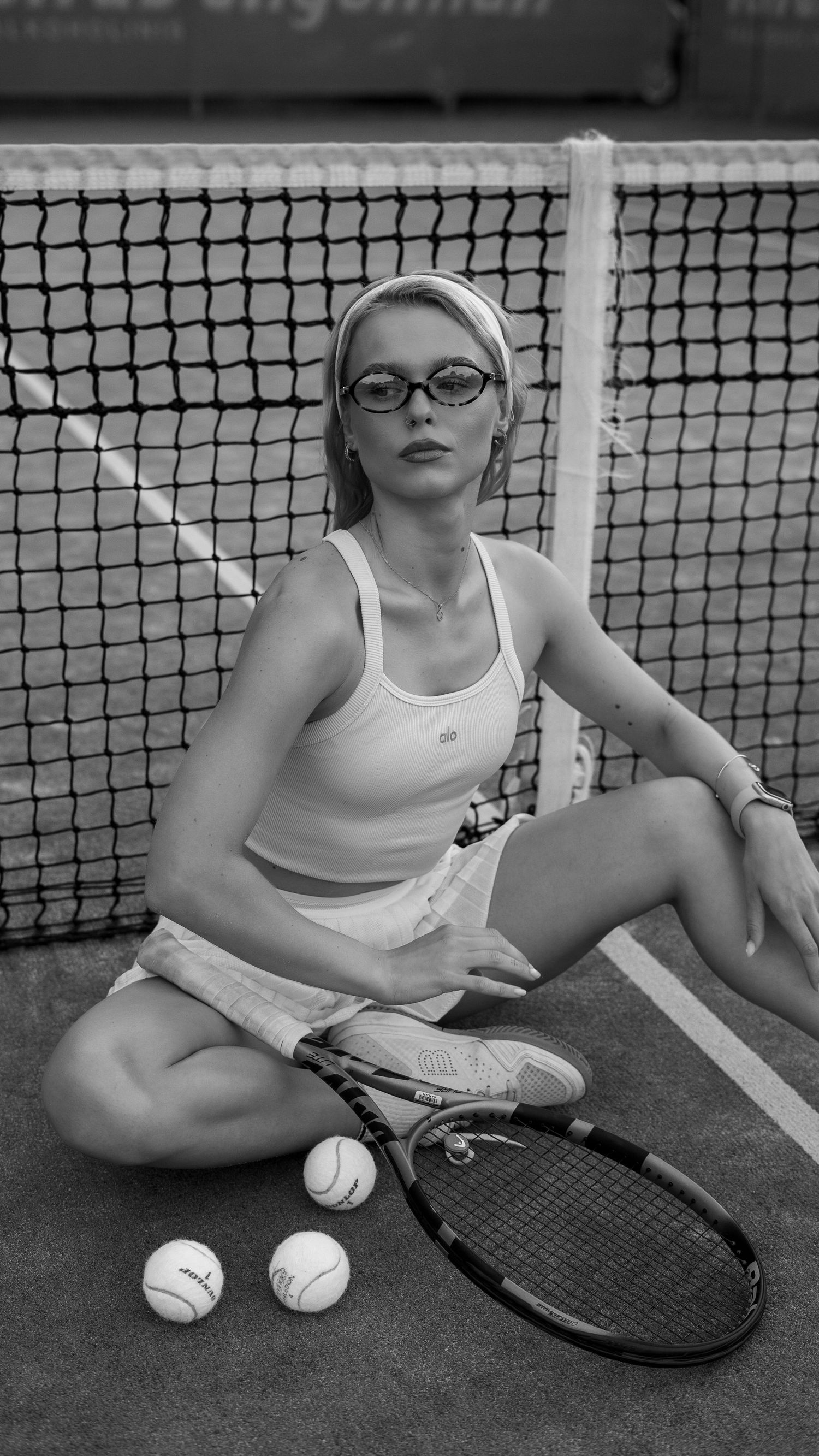 Blonde woman in white tennis outfit sitting by net with racket and balls.