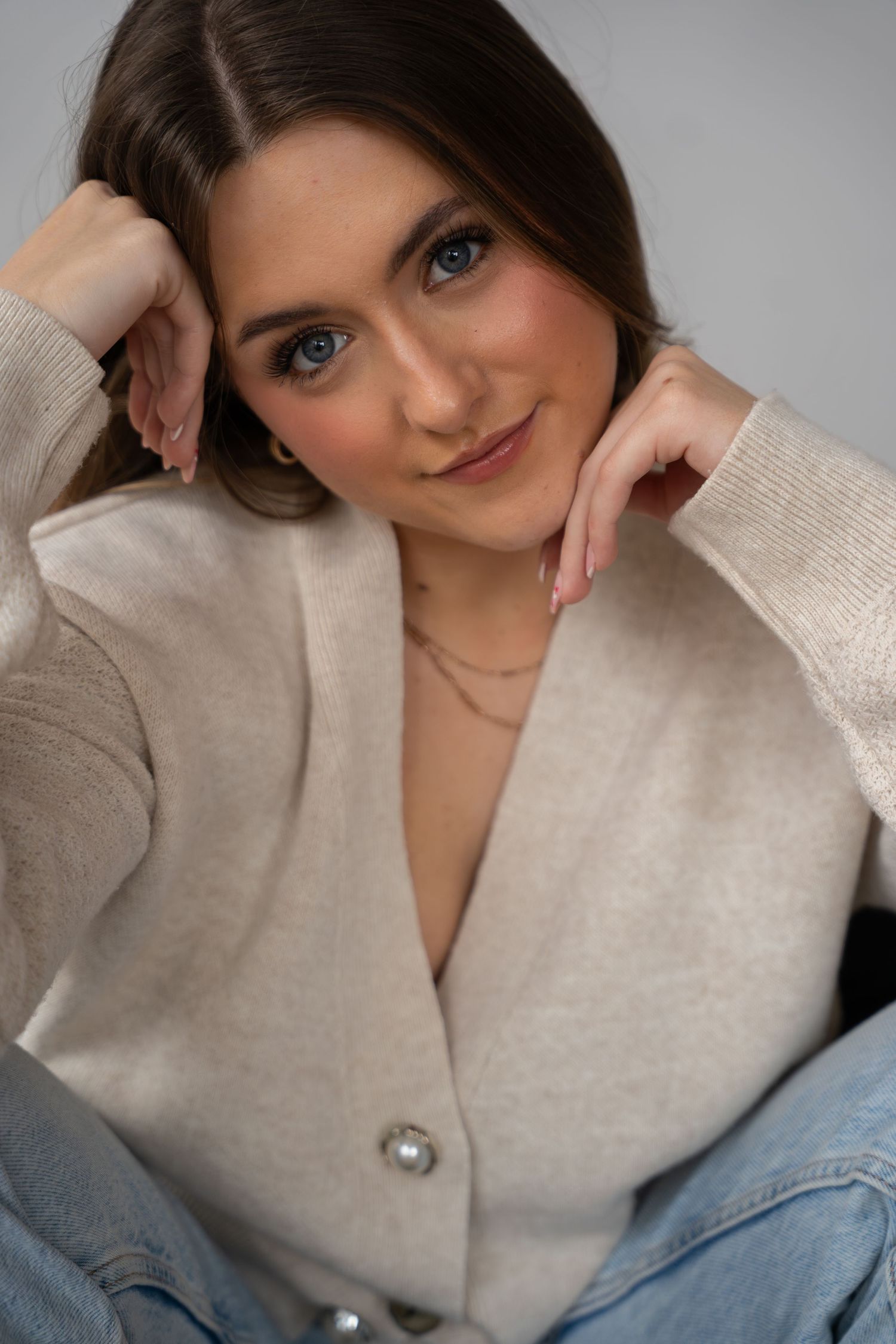Close-up portrait of blue-eyed brunette woman in beige cardigan smiling with hands framing her face.