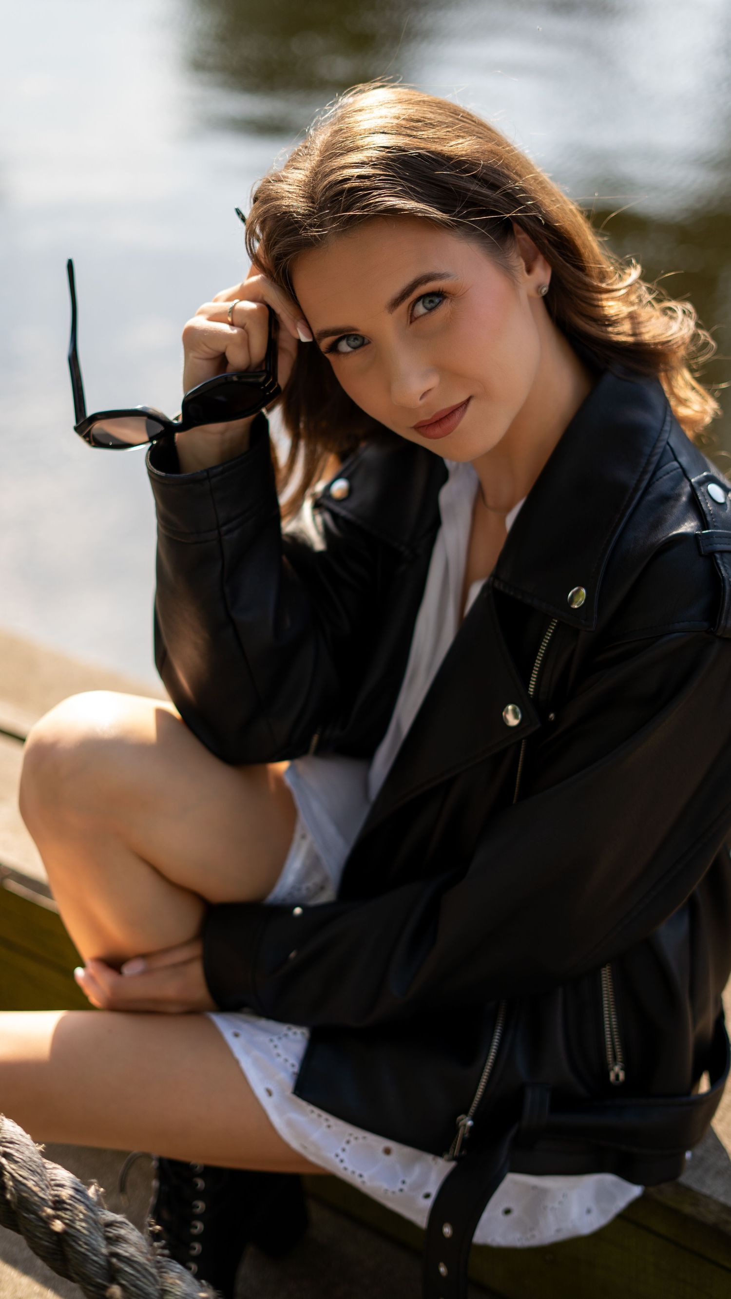 Woman in black leather jacket and white dress sitting outdoors holding sunglasses near water in sunlight.