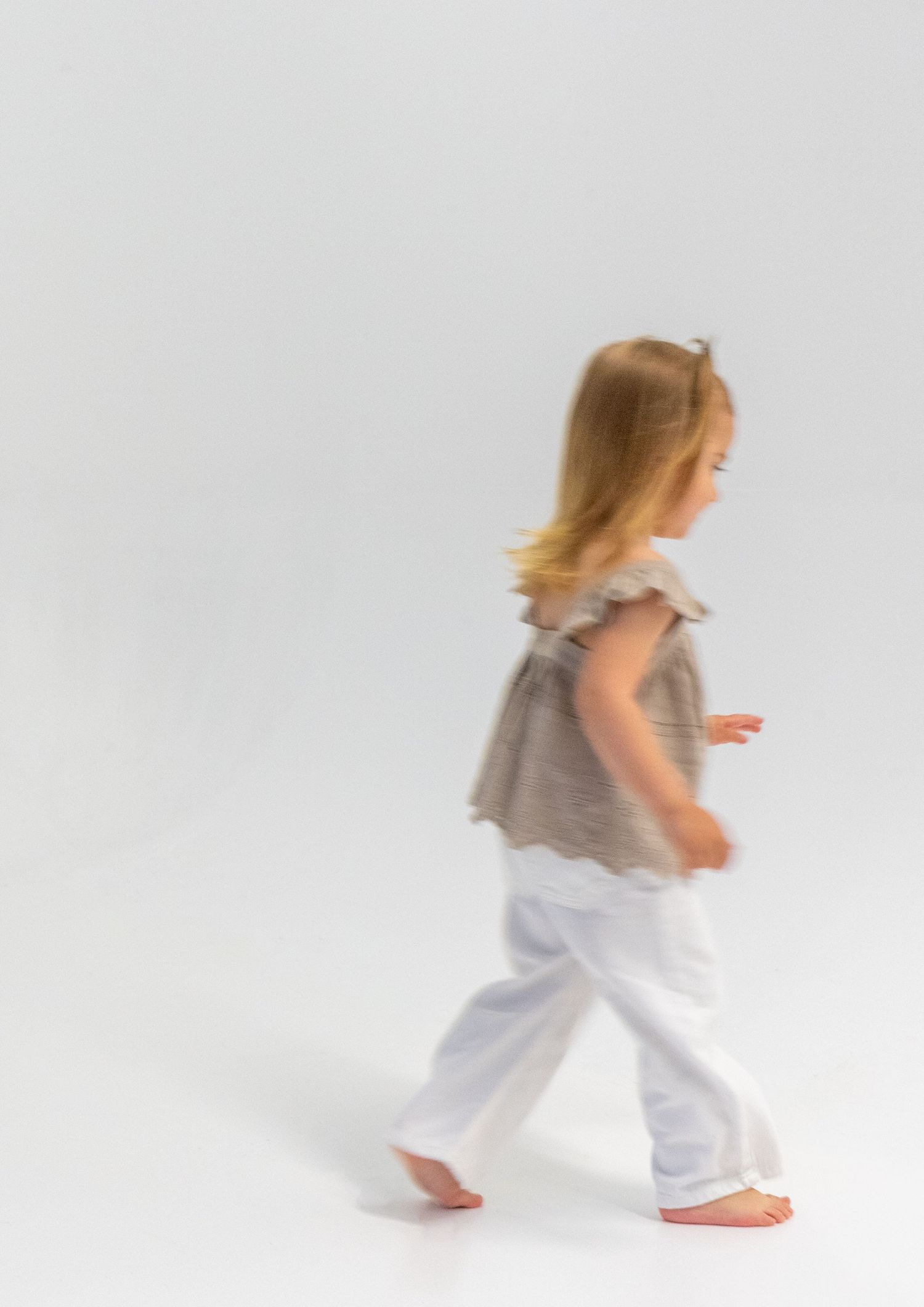 Blurred motion shot of young blonde girl walking in white pants and gray ruffled top on white background.