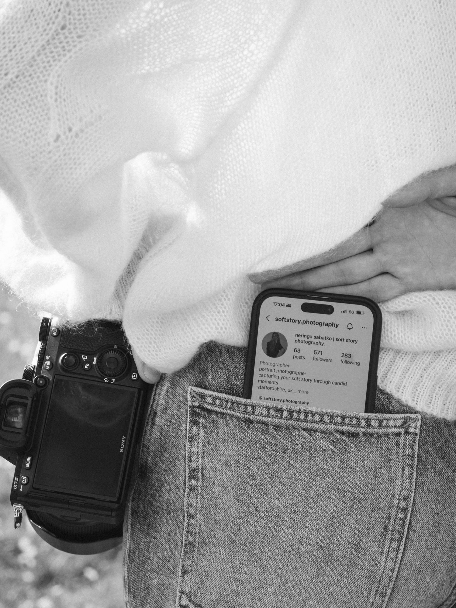 Black and white photo of person holding camera with phone showing Instagram profile in jeans pocket.