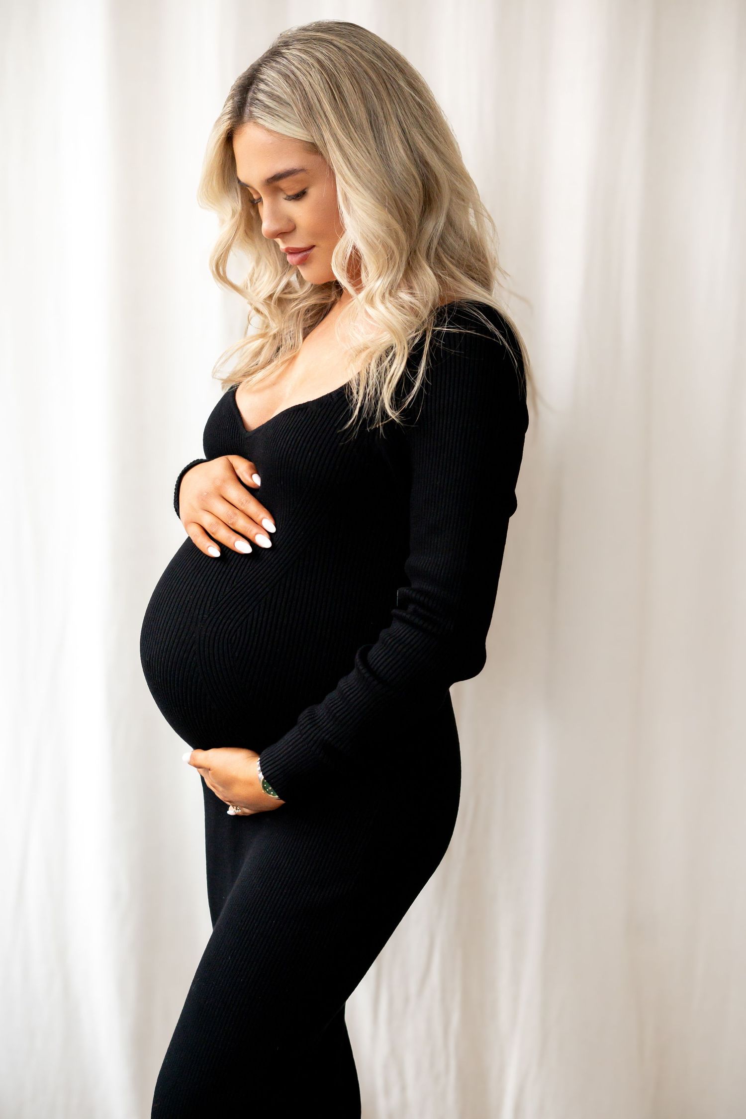 Pregnant woman in black ribbed long-sleeve dress looking down tenderly at her baby bump.