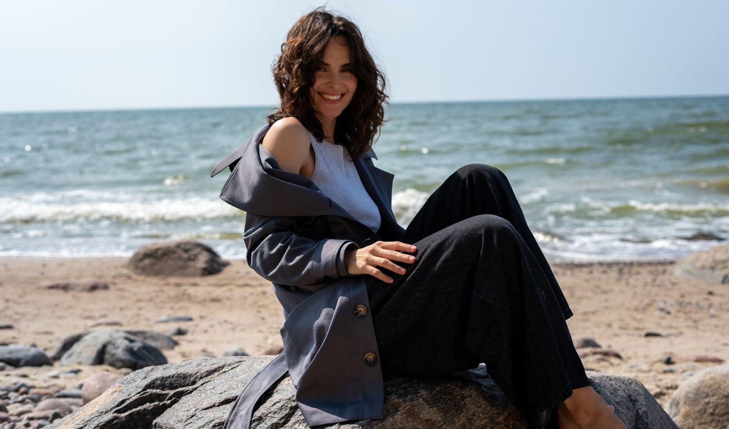 Smiling woman in gray and navy outfit relaxing on beach rocks with sunny ocean backdrop.