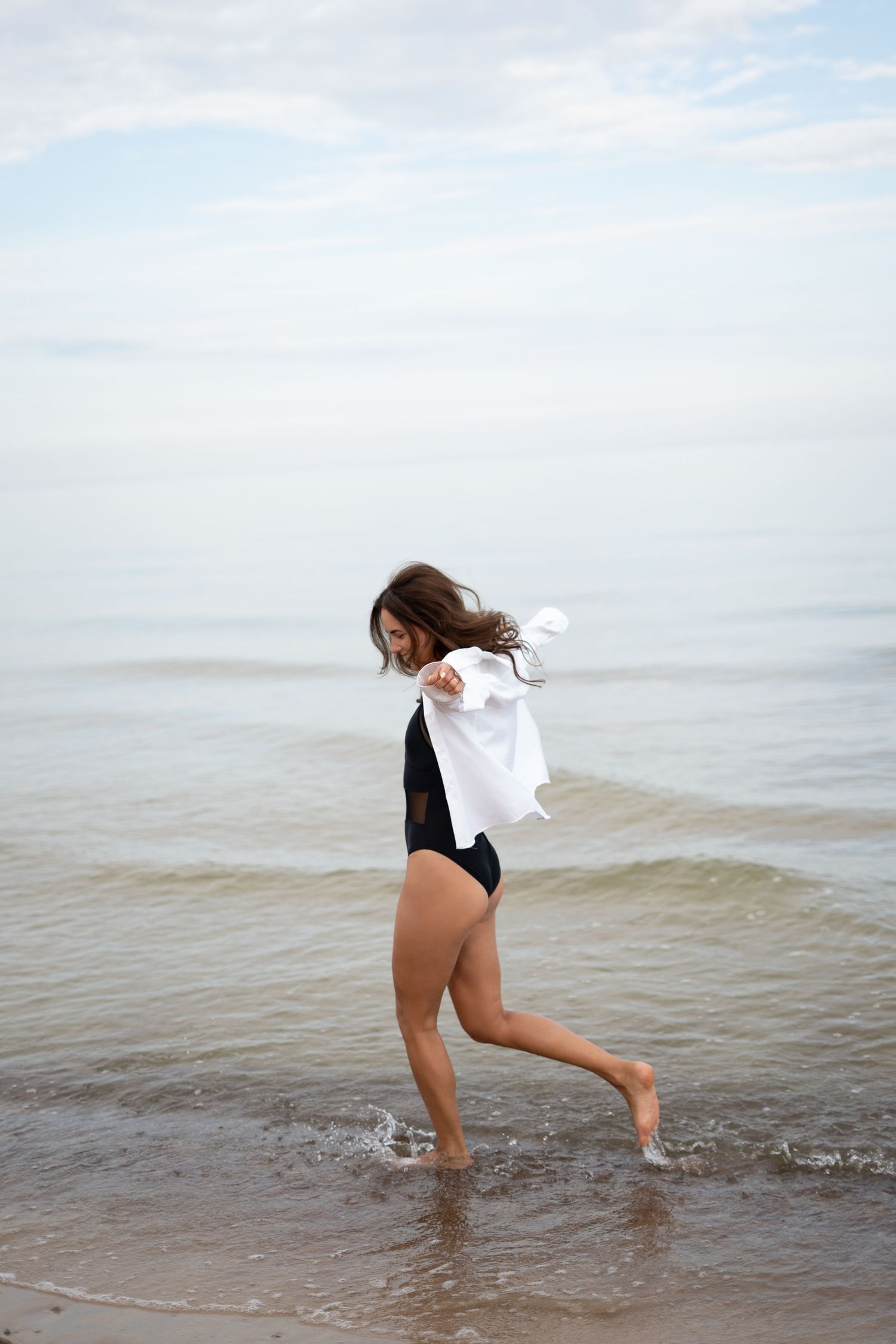 Woman in black swimsuit holding white shirt walking through shallow ocean waves on beach.
