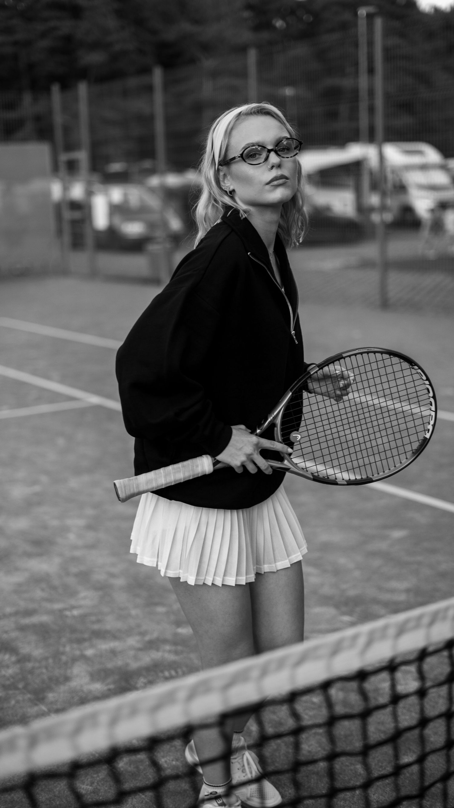 Blonde woman in black jacket and white skirt holding tennis racket on court.
