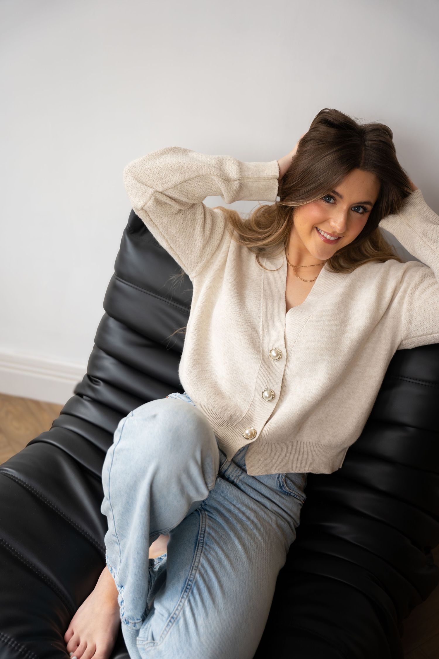 Smiling woman in cream cardigan and blue jeans sitting relaxed on a black leather bean bag chair.