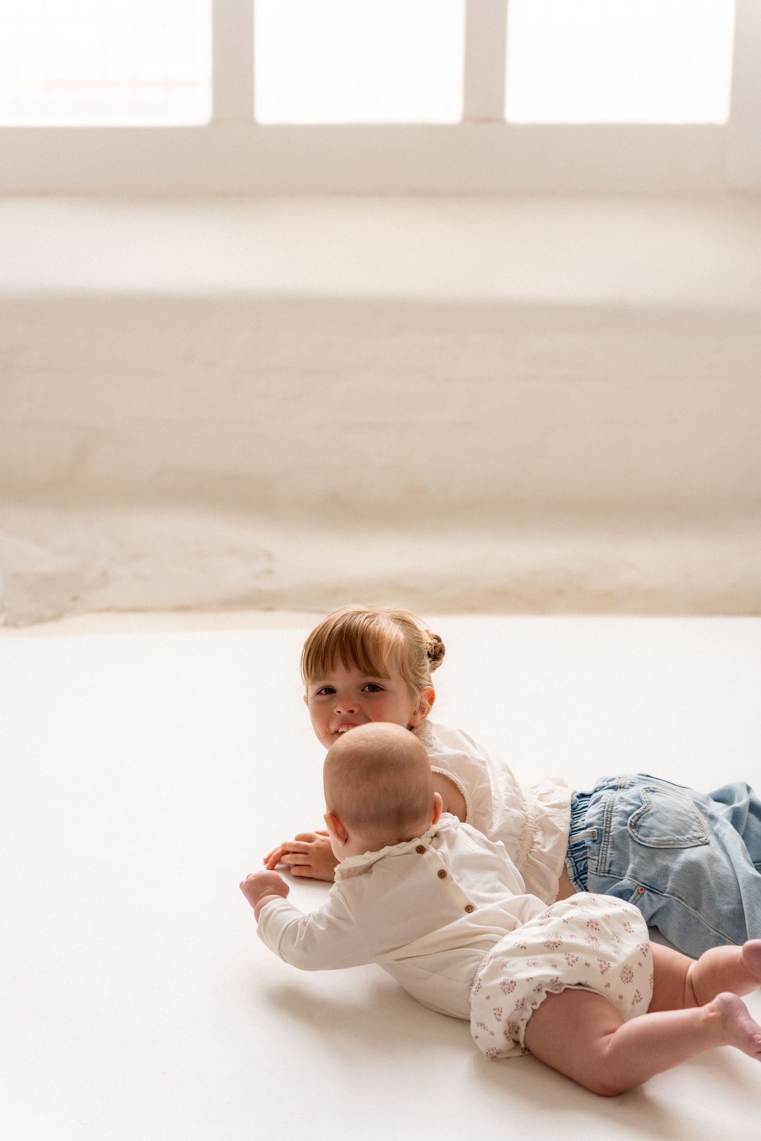 Two young siblings, a toddler girl and baby, playing together on a white floor in a bright room.