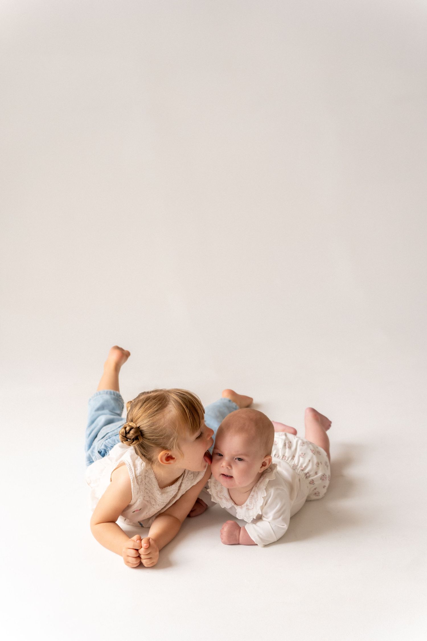 A toddler girl in blue jeans kisses a baby sibling while both lie on a white studio floor.