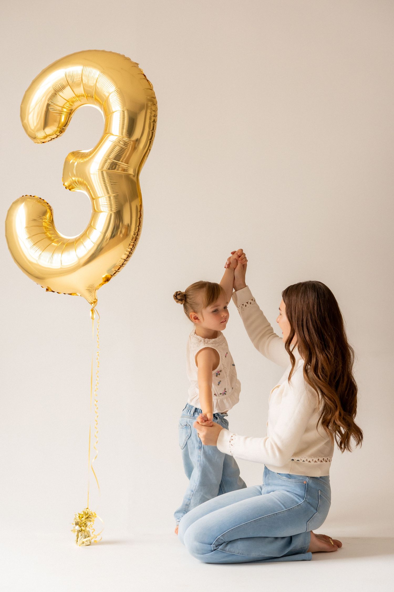 Mother kneeling with toddler daughter beside a gold number 3 balloon on white background for 3rd birthday celebration.