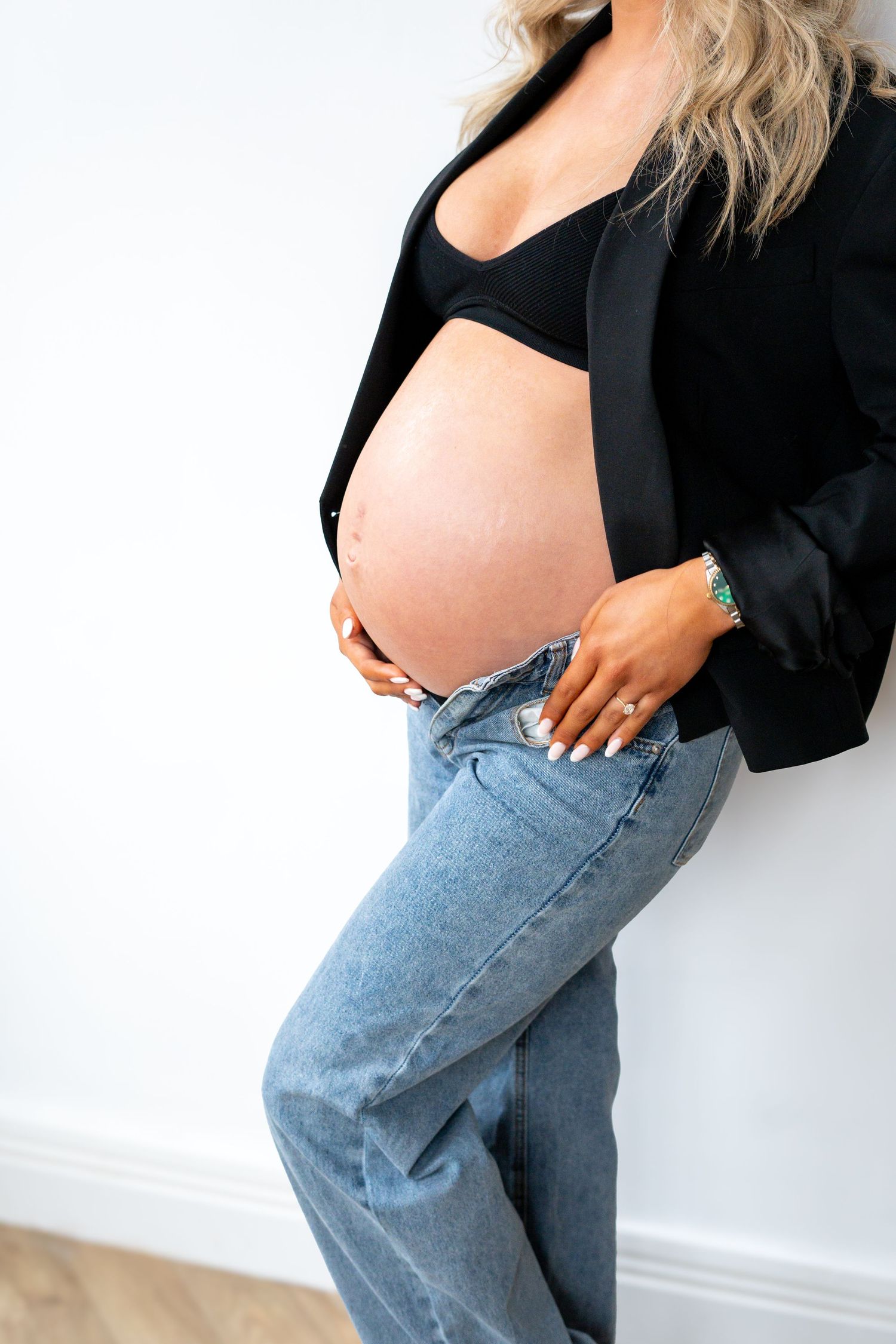 Close-up of pregnant woman in black blazer and jeans leaning against white wall, cradling bare bump.