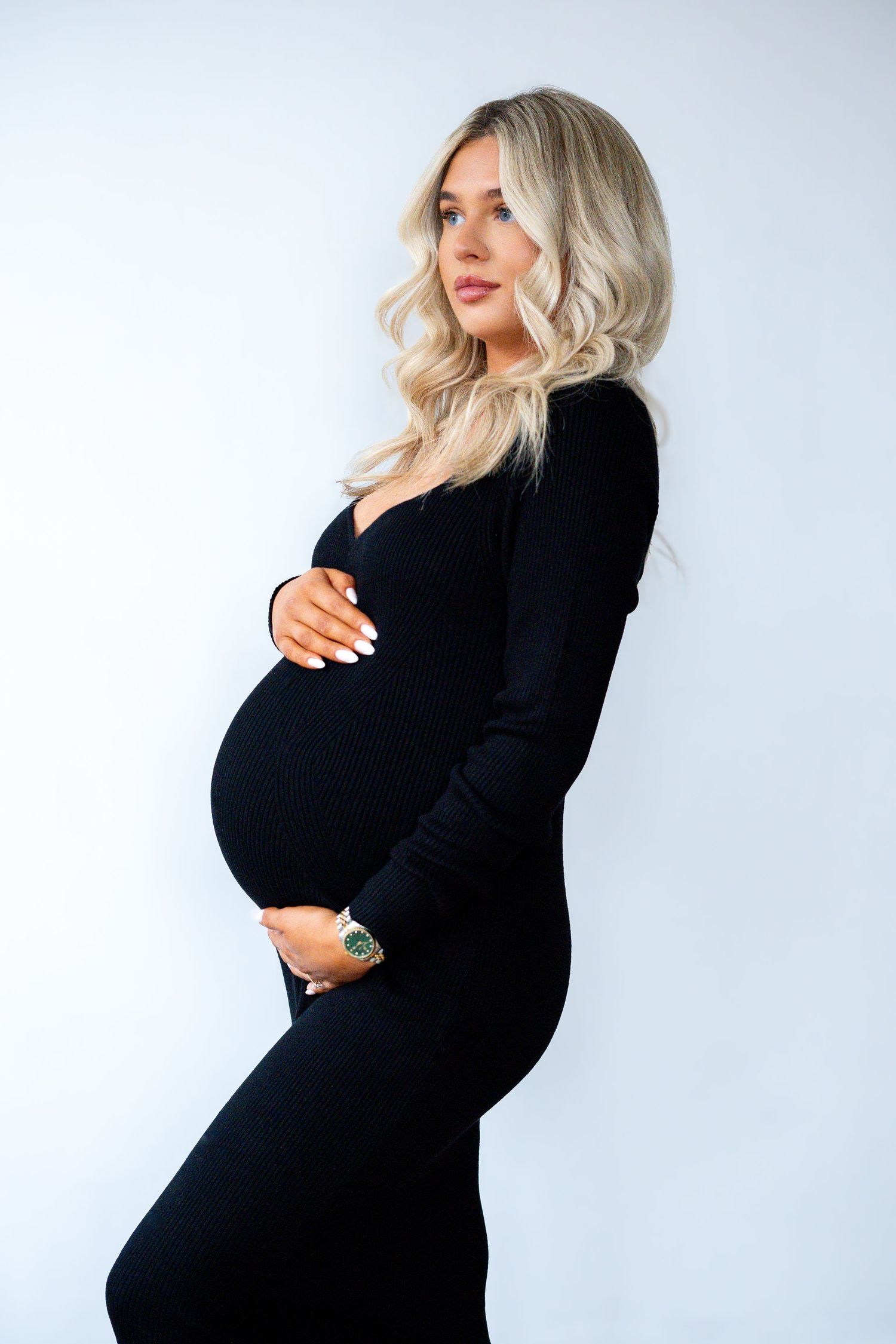 Side view of smiling pregnant woman in black fitted dress wearing green watch, holding bump.