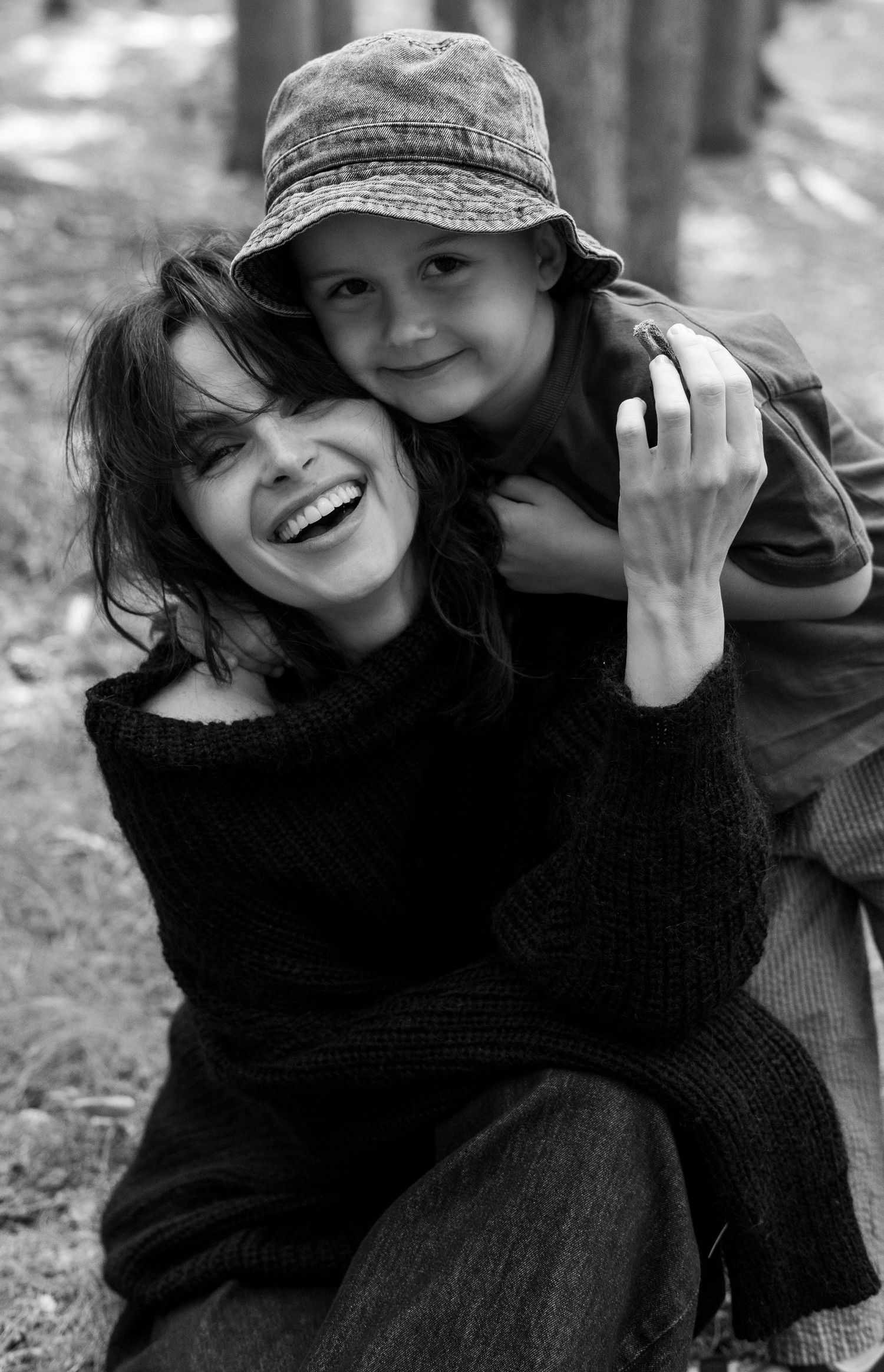 Black and white portrait of smiling woman with young child hugging her from behind outdoors.