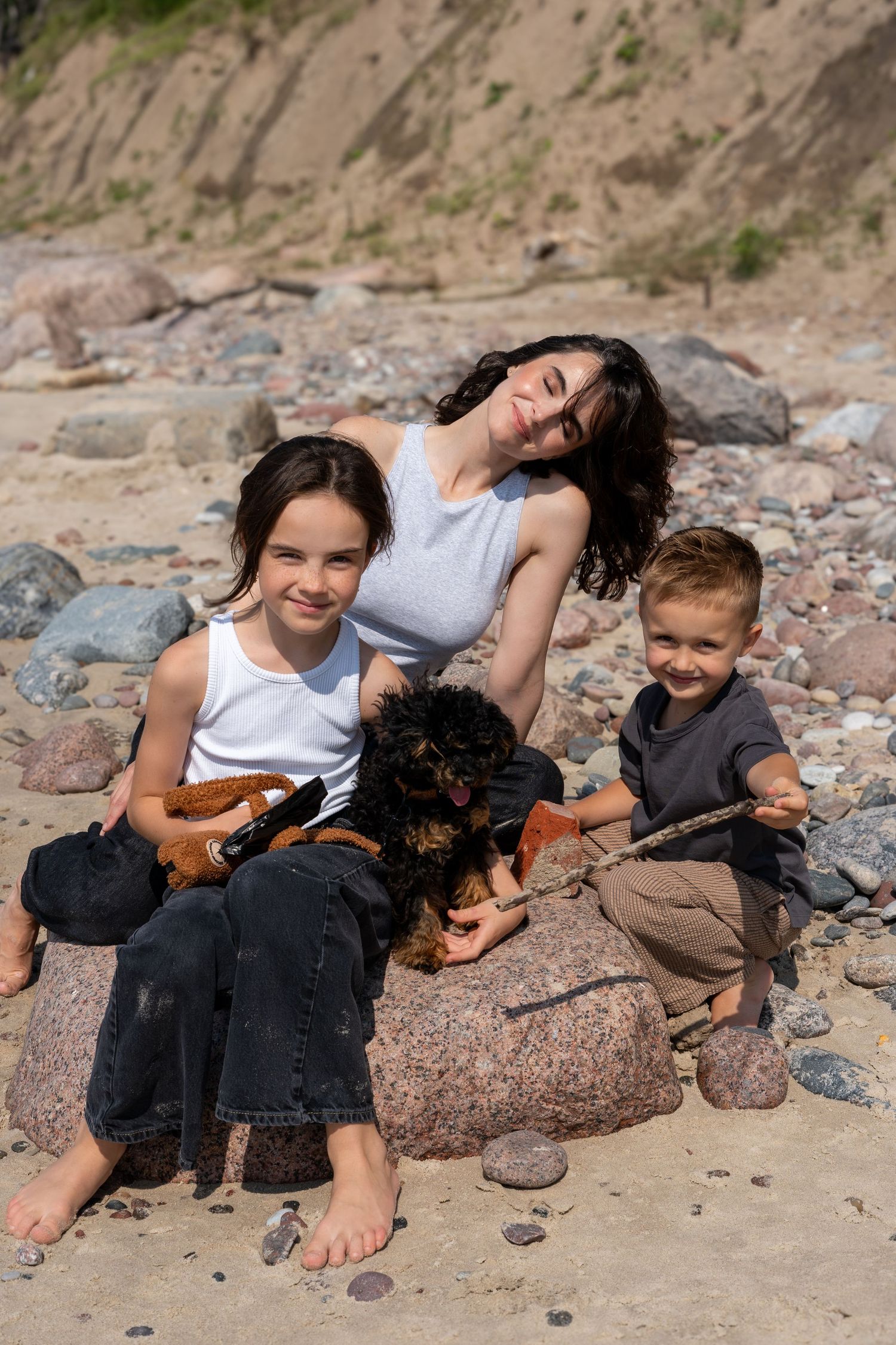 A mother and two children sit on rocks at a beach with a black dog on a sunny day.