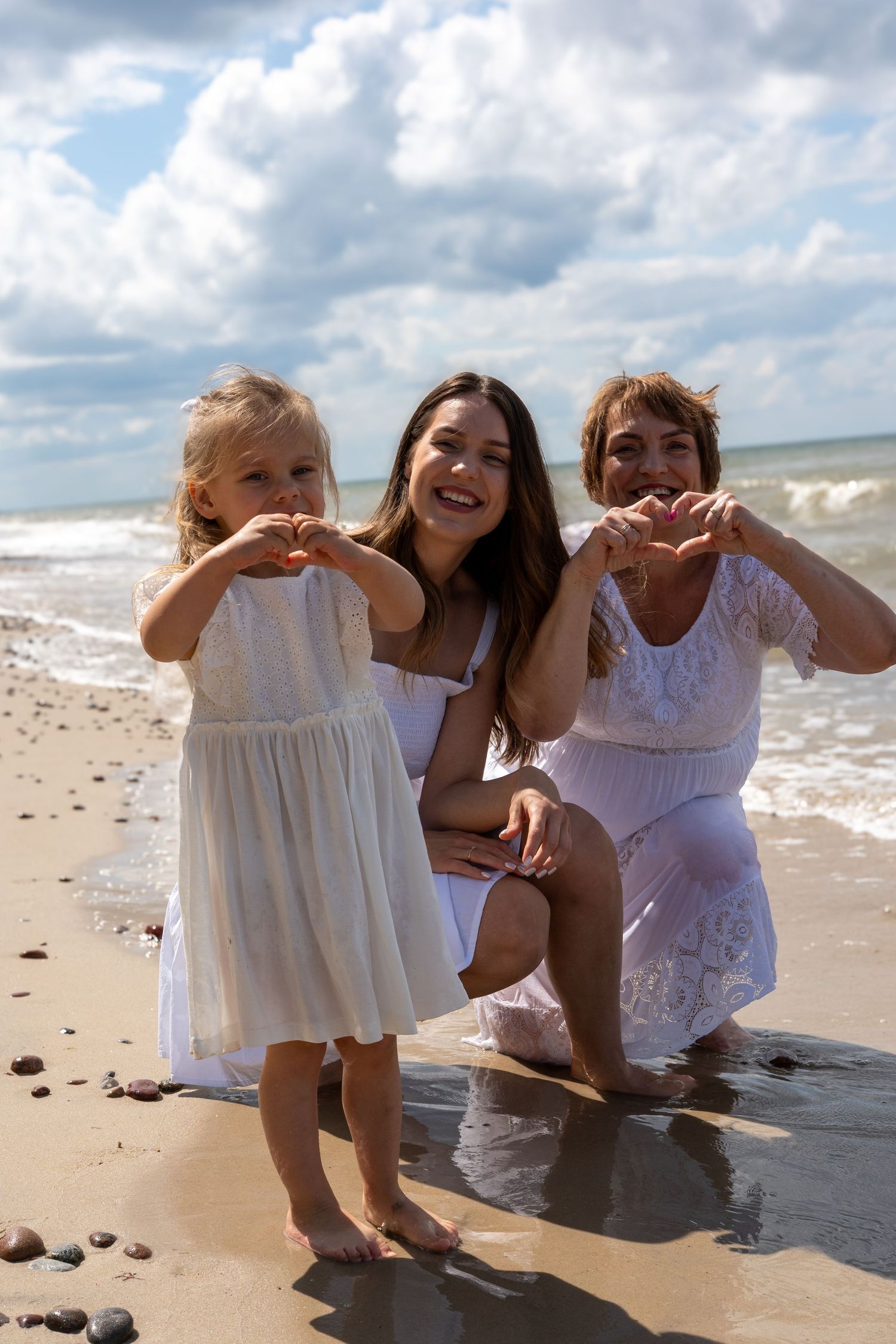 Three generations of women in white dresses make heart shapes with hands on a sunny beach.