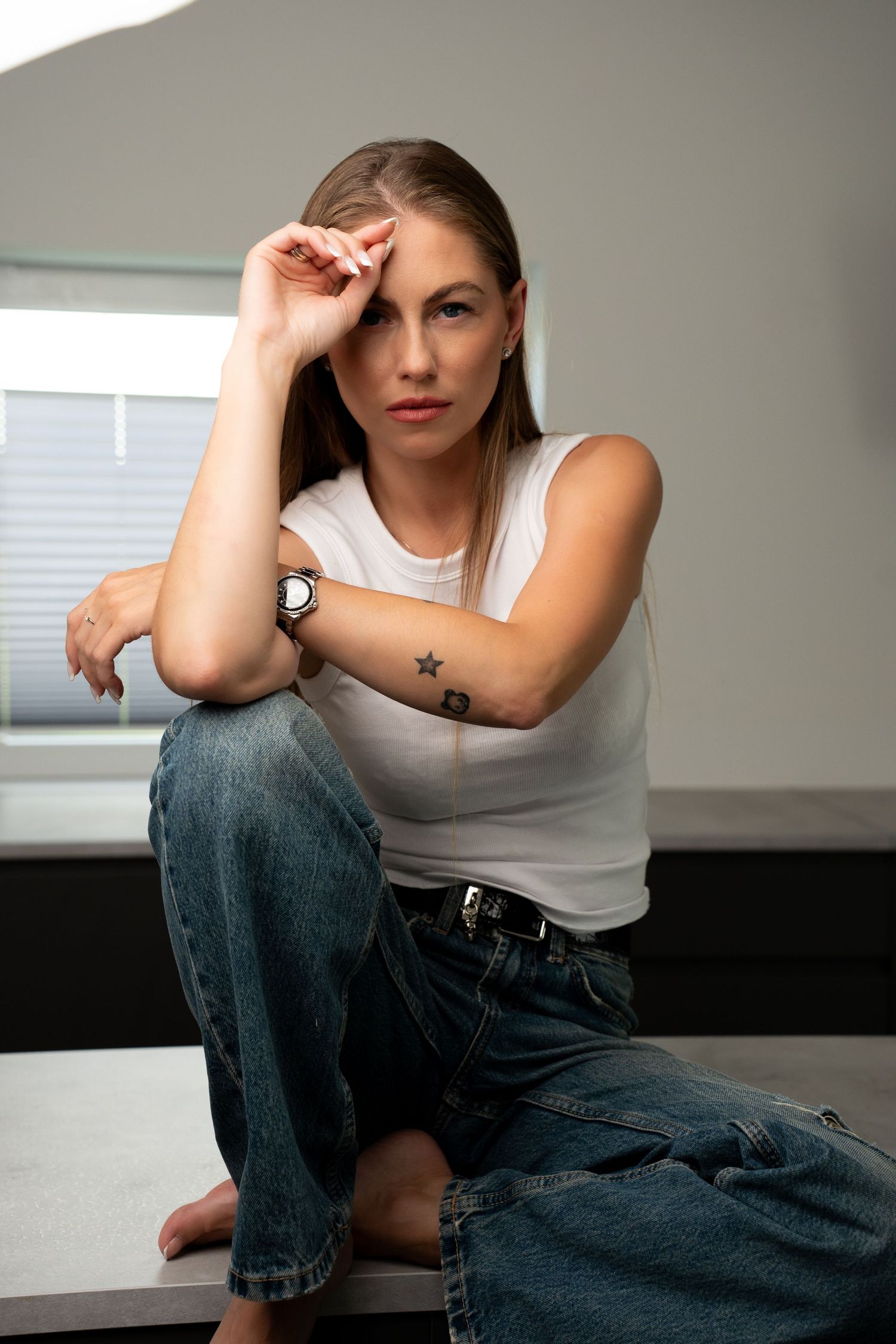 Woman with tattoos sits on surface near window wearing white sleeveless top, blue jeans, and a watch.