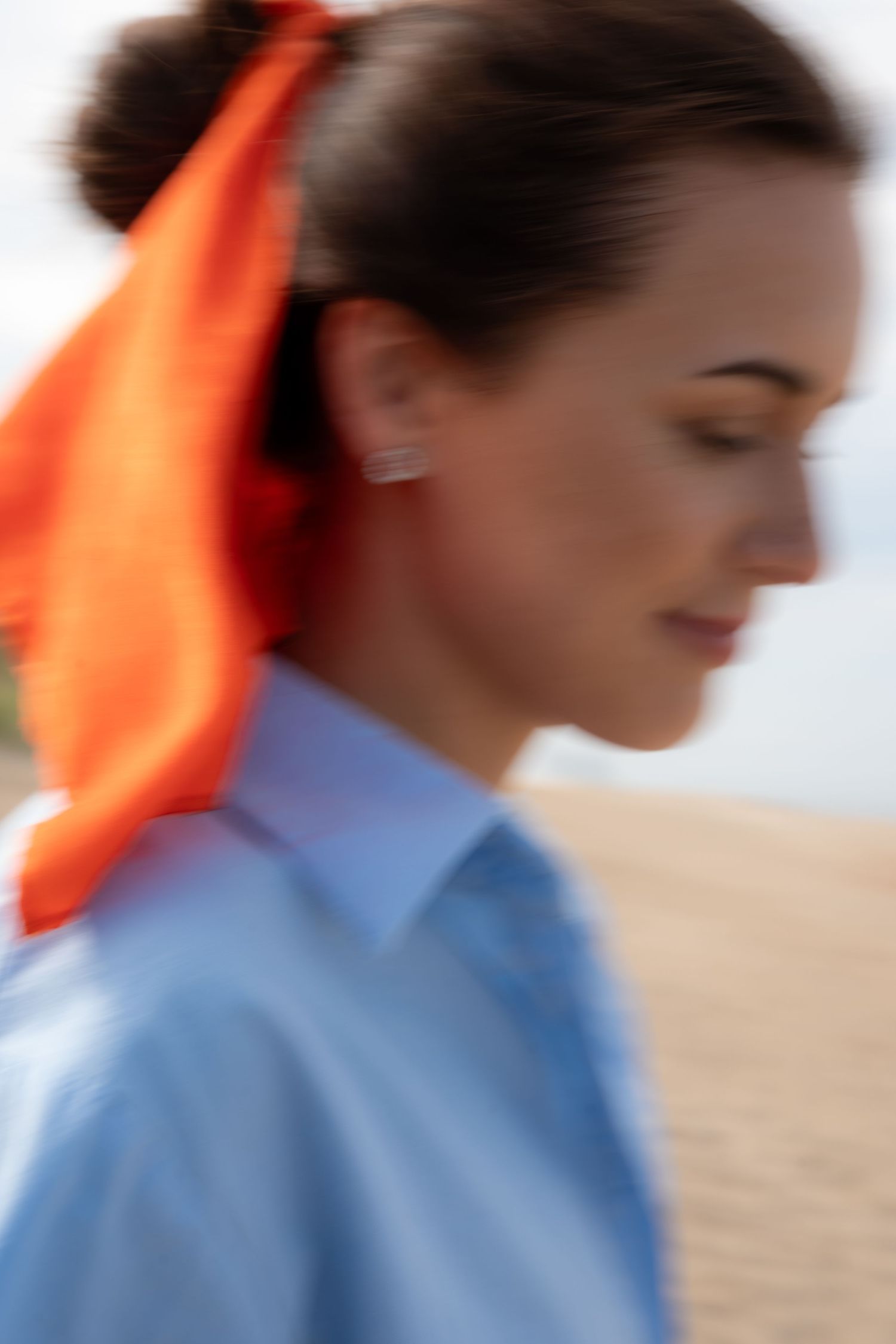 Blurred close-up of brunette woman with orange hair scarf and blue shirt on sandy beach.
