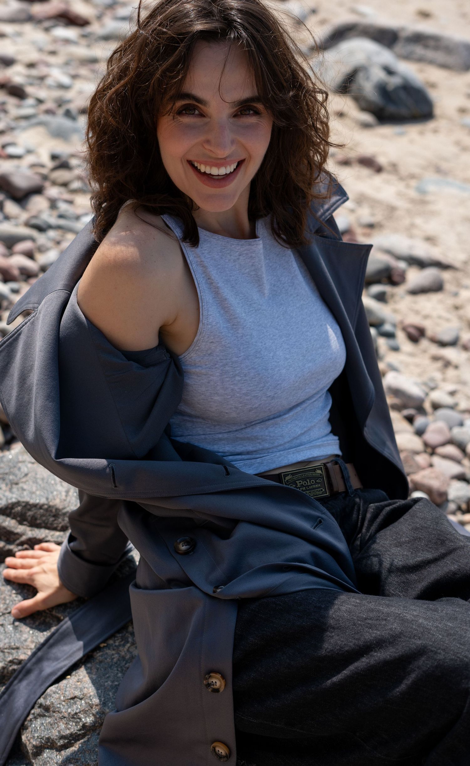 Laughing woman in white tank top and dark jacket sitting on pebble beach in sunlight.