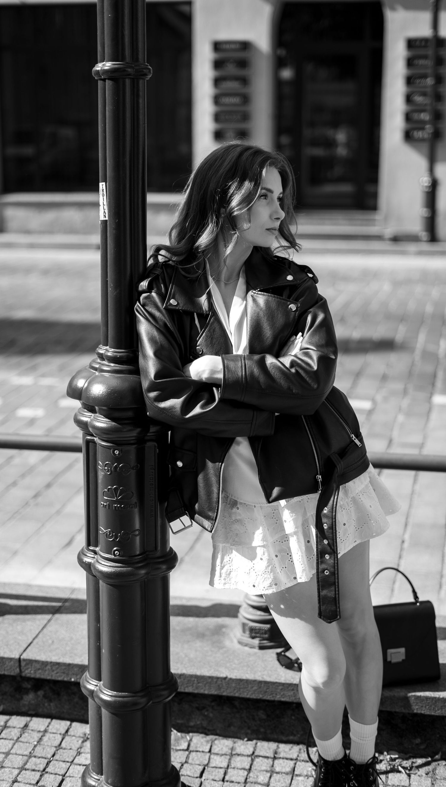 Stylish woman in black leather jacket and white dress leaning on street lamp in urban black and white photo.