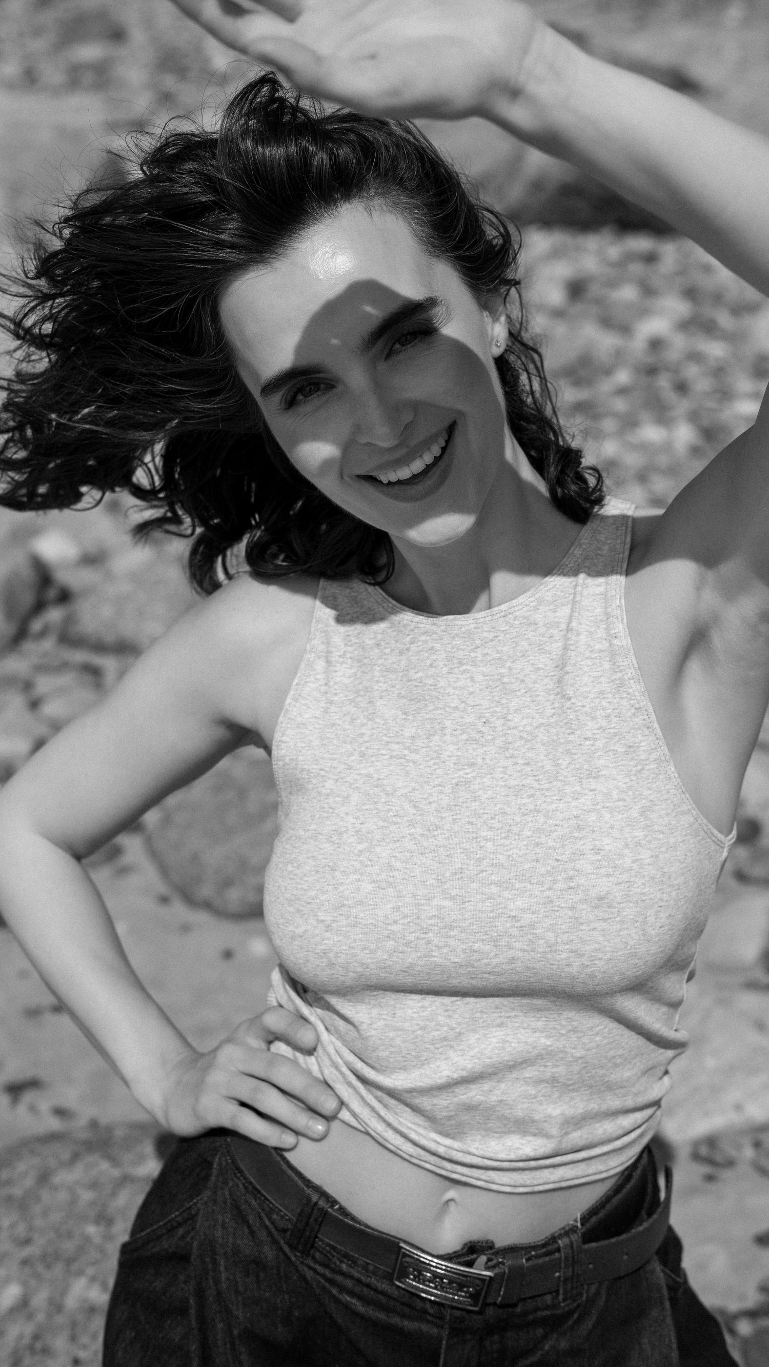 Black and white photo of smiling woman in crop top with arms raised, curly hair blowing in breeze.