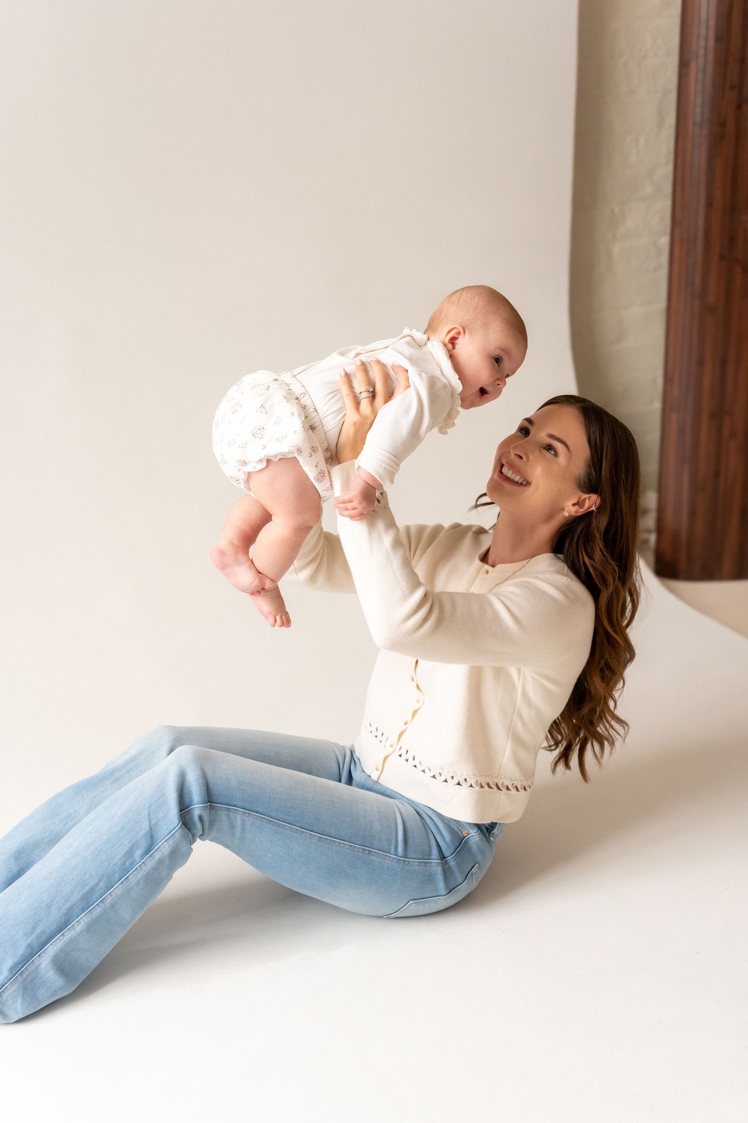 A smiling mother sits on the floor lifting her happy baby in the air, both dressed in white in a bright studio.