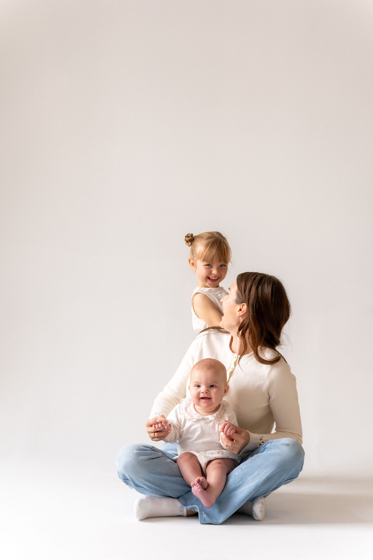 Mother sitting cross-legged with baby in lap and toddler daughter perched on her shoulder on white background.