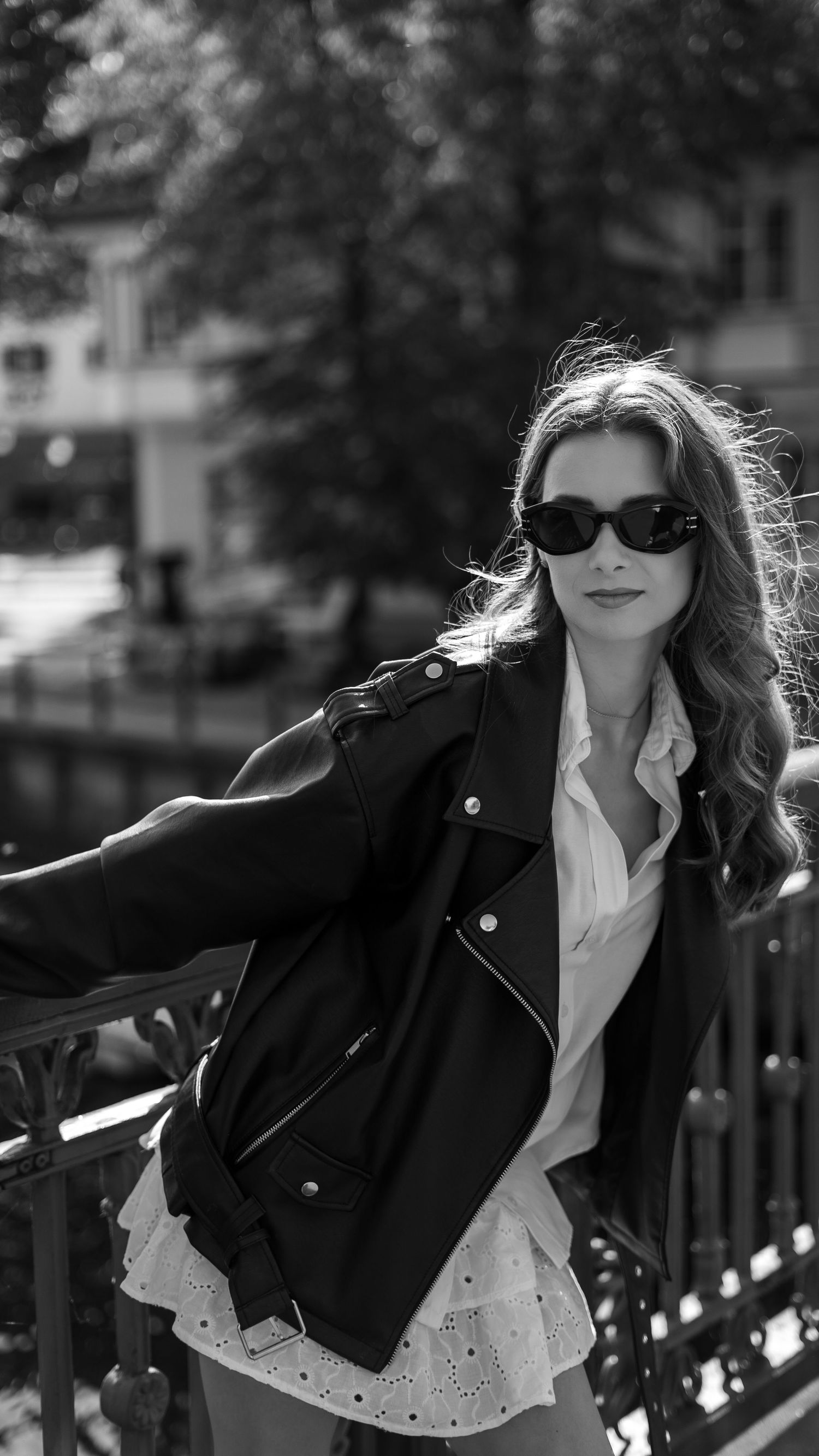 Woman wearing black sunglasses and leather biker jacket over white dress posing on bridge in black and white.