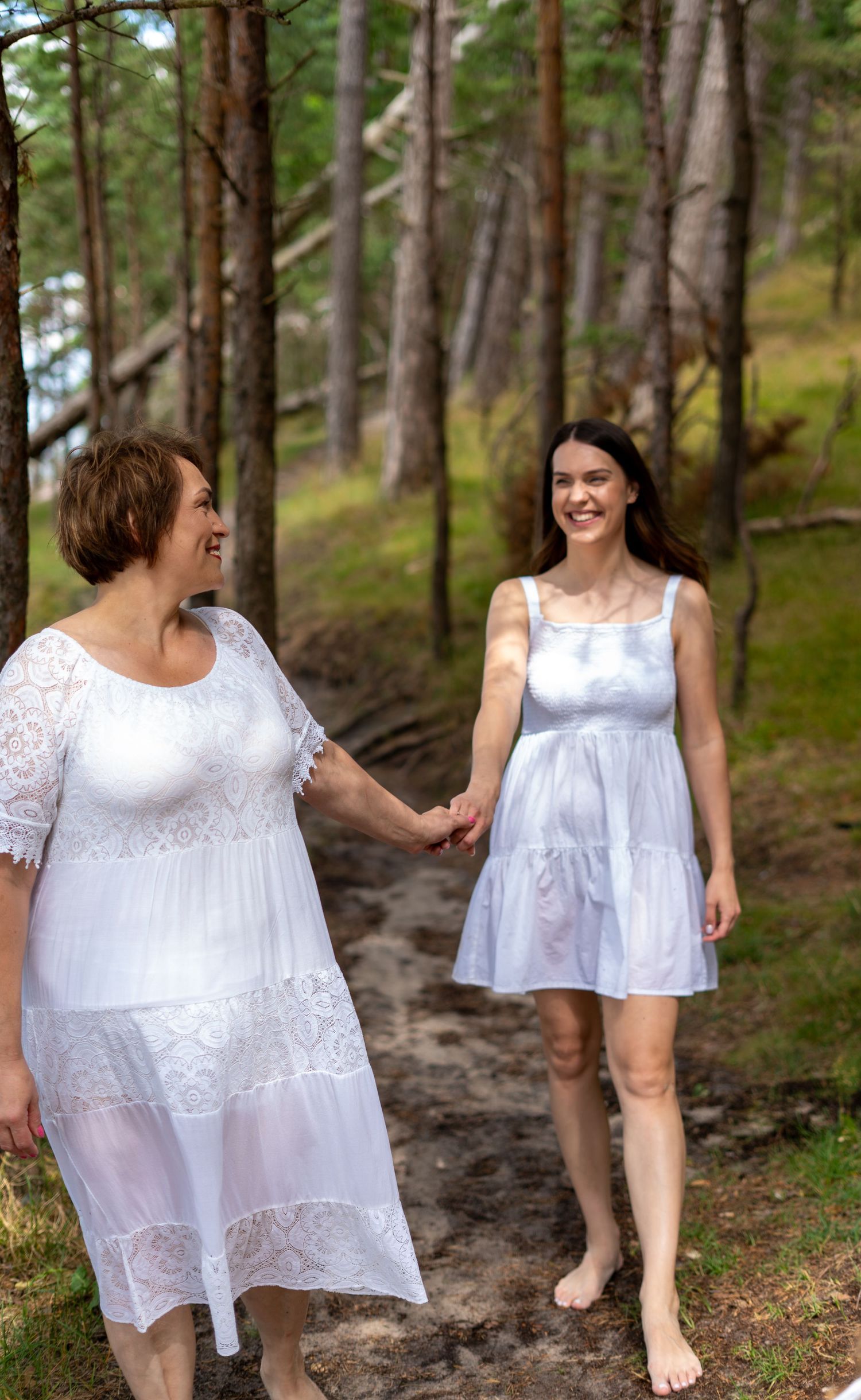 Two women in white dresses hold hands while walking barefoot on a sandy forest trail.